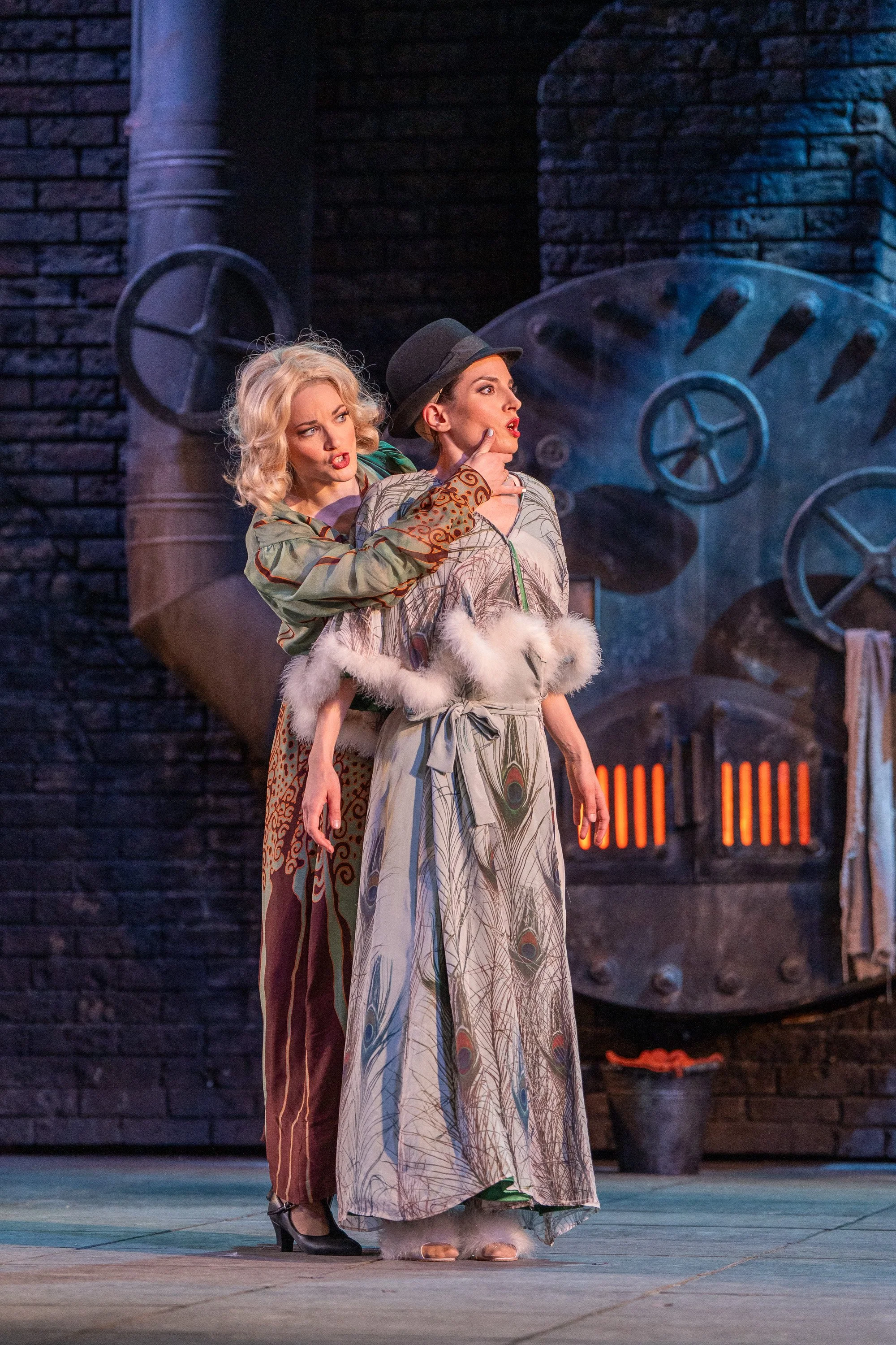 Two women in theatrical costumes are performing on stage with a brick wall and industrial backdrop. The woman on the left has curly blonde hair in a vintage style, wearing patterned wide-leg pants and a blouse, and is leaning on the other woman's shoulder, looking at her with a concerned expression. The woman on the right has straight hair, wearing a hat, a long patterned dress, and fluffy footwear, looking away with a dramatic expression. The scene suggests a theatrical or dance performance.