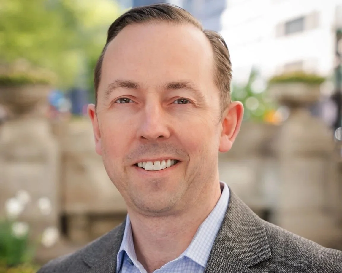 Close-up portrait of Simon Barcoe, the founder of Klaracon, he is smiling man with short brown hair, wearing a gray blazer and a light blue collared shirt, outdoors with blurred background including greenery and buildings.