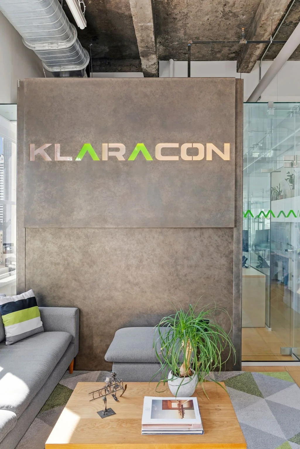 A modern office lobby with a concrete wall featuring lit-up sign that reads "KLARACON" in green and white letters. There are gray sofas, a wooden coffee table with a book, a decorative sculpture, and a potted plant. Glass walls reveal additional office space inside.