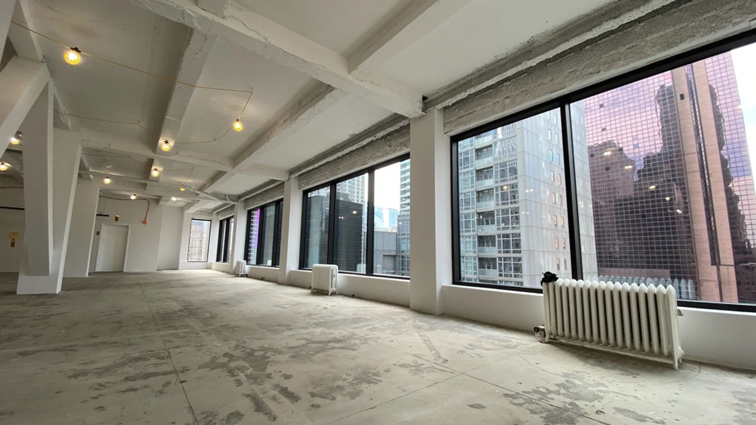 An empty, unfinished high-rise room with large floor-to-ceiling windows showing city buildings outside. The ceiling has exposed beams and hanging lights, and the floor is concrete.