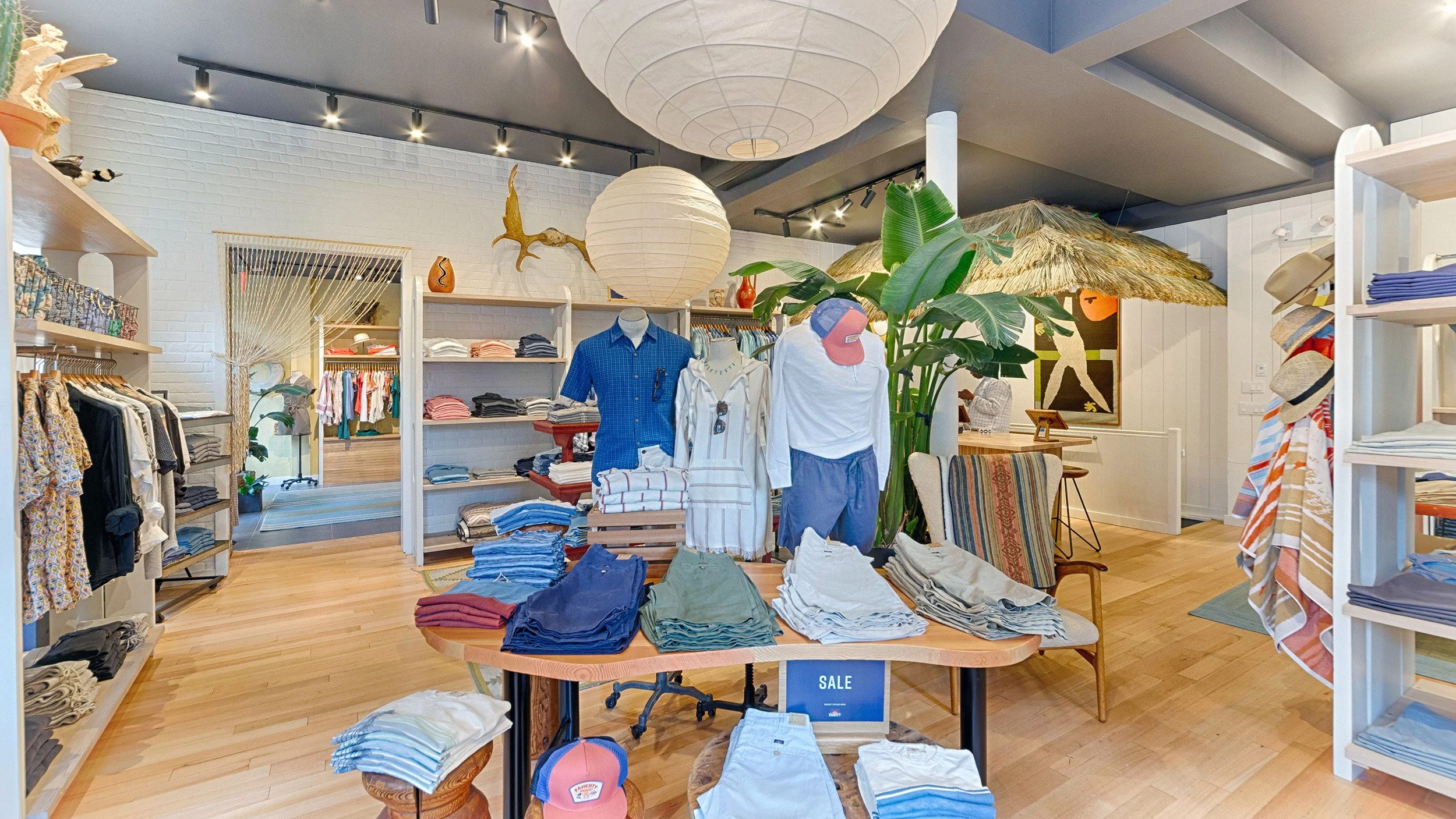 P670 by Klaracon - a retail clothing store with a variety of racks and table displays with casual and tailored casual clothing about, decor is beachy and light and airy with painted white brick walls and large paper lantern lights