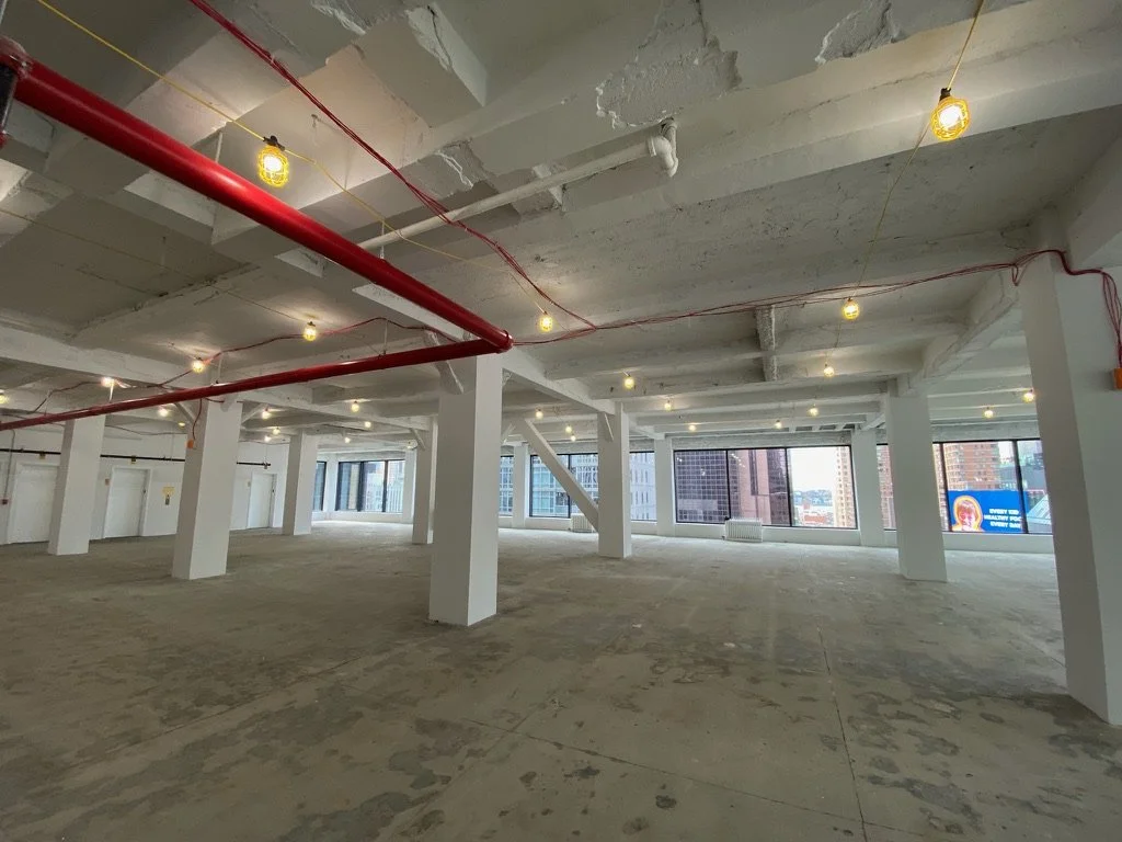 Urban interior space of a white box industrial space with white columns and red pipe running along the ceiling and visible large windows looking out onto an urban landscape