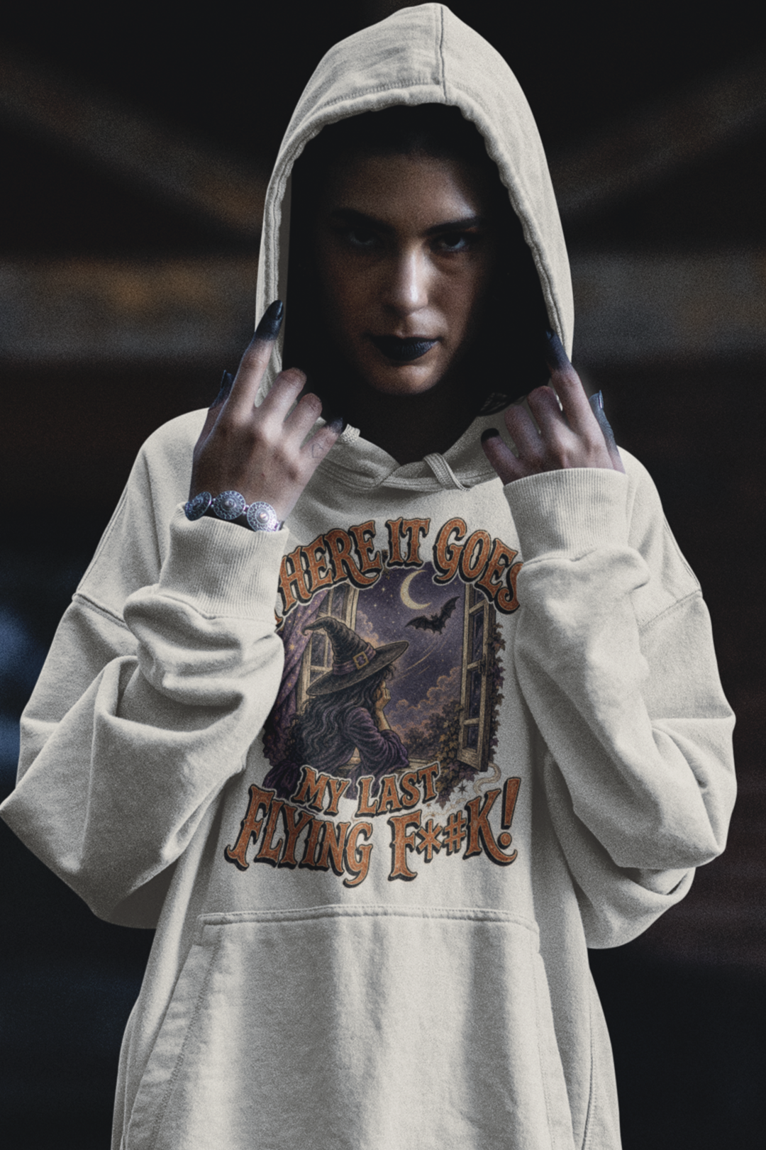 A woman wearing a light-colored hoodie with a graphic and text on it, dark lipstick, and dark painted nails, looking directly at the camera with a serious expression and adjusting her hood.