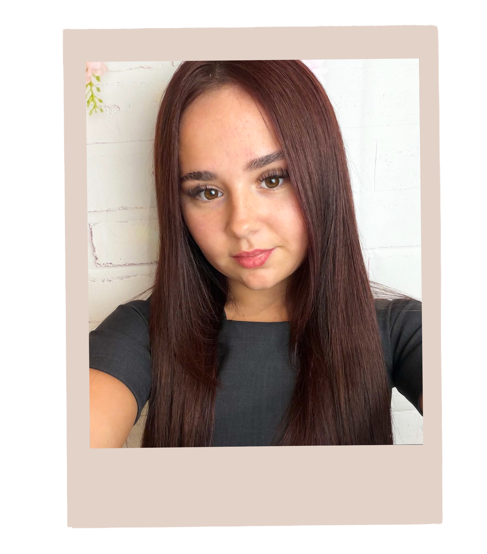 Close-up of a young woman with straight, long, reddish-brown hair and light brown eyes, wearing a black top, standing in front of a white wall with visible bricks.