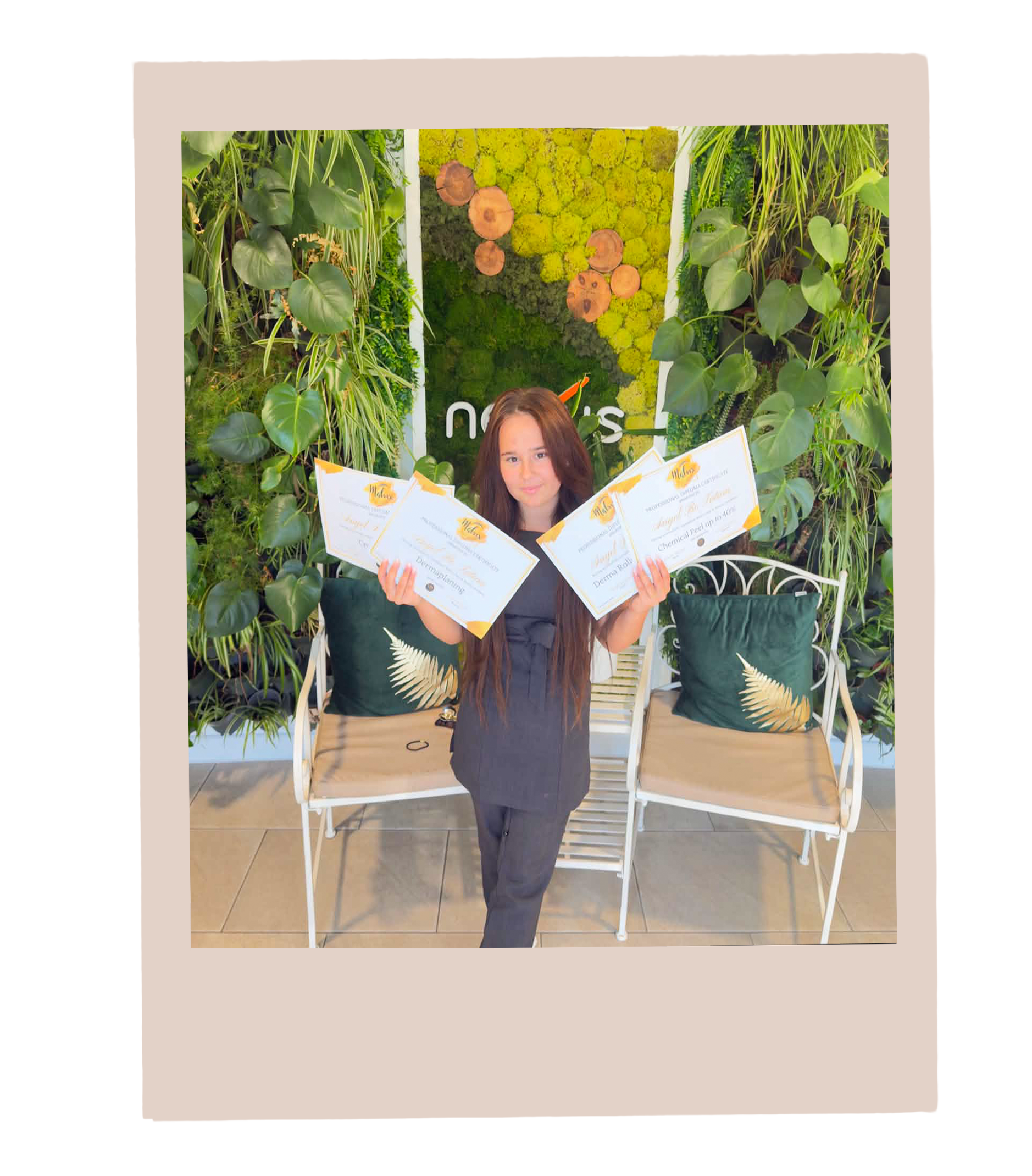 A young girl with long brown hair holds four certificates in front of her. She stands in front of a decorative green wall with plants on either side, and there are two white chairs with black cushions behind her.