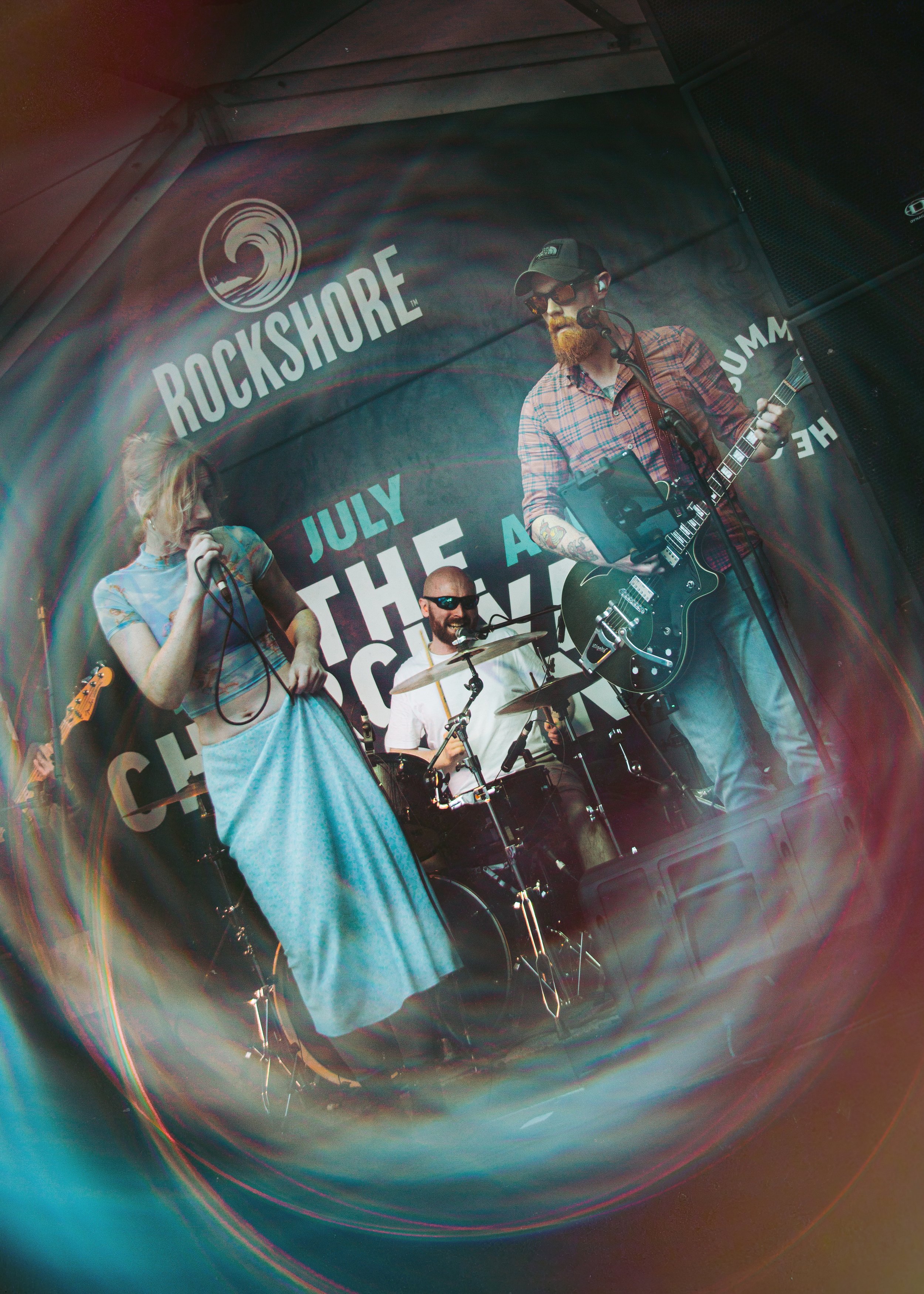 A band performing on stage at Rockshore music event with a female singer, a drummer, and a guitarist. The backdrop displays the event name and date.