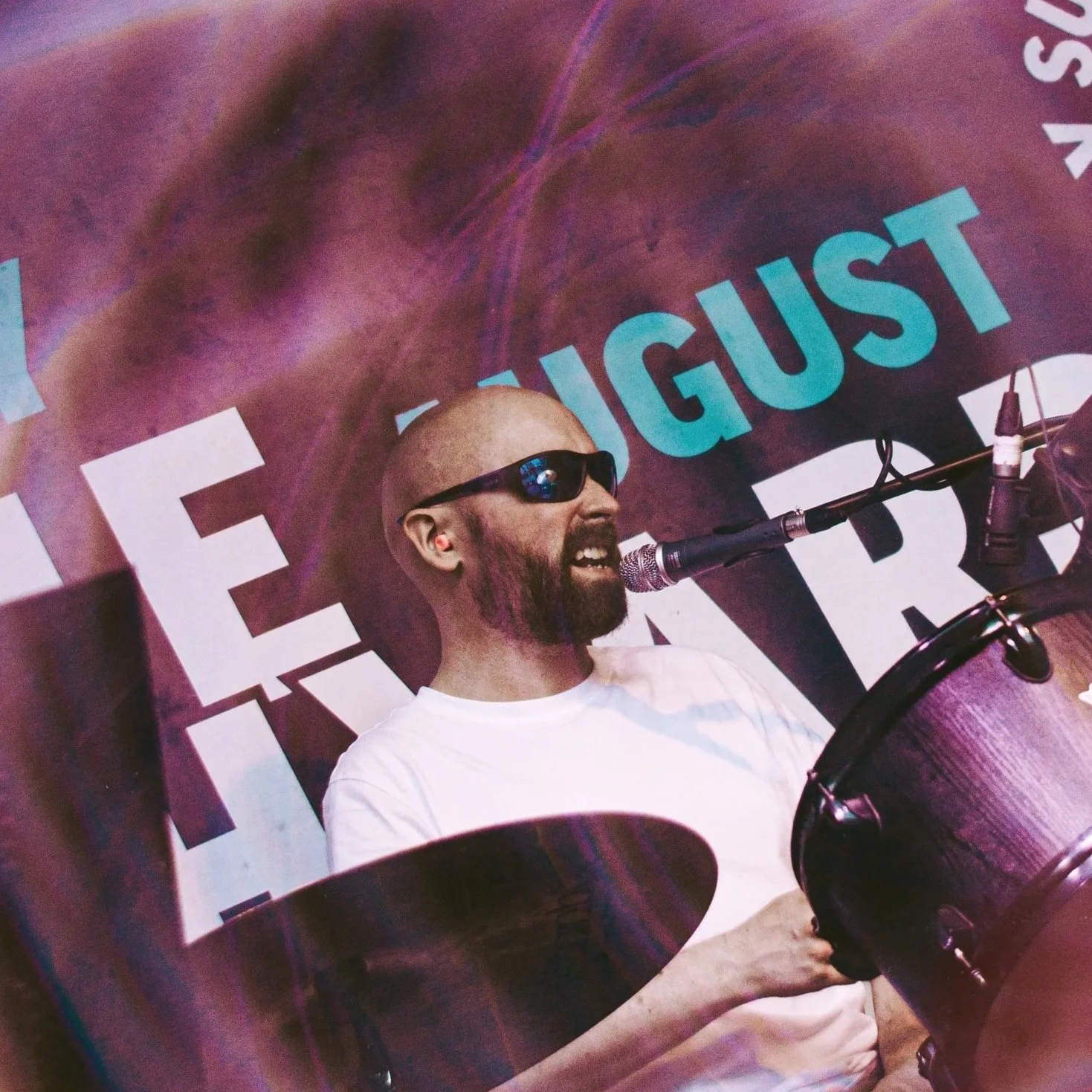 Man with a beard and sunglasses playing drums in front of a microphone, with a large purple banner behind him that reads 'AUGUST'.