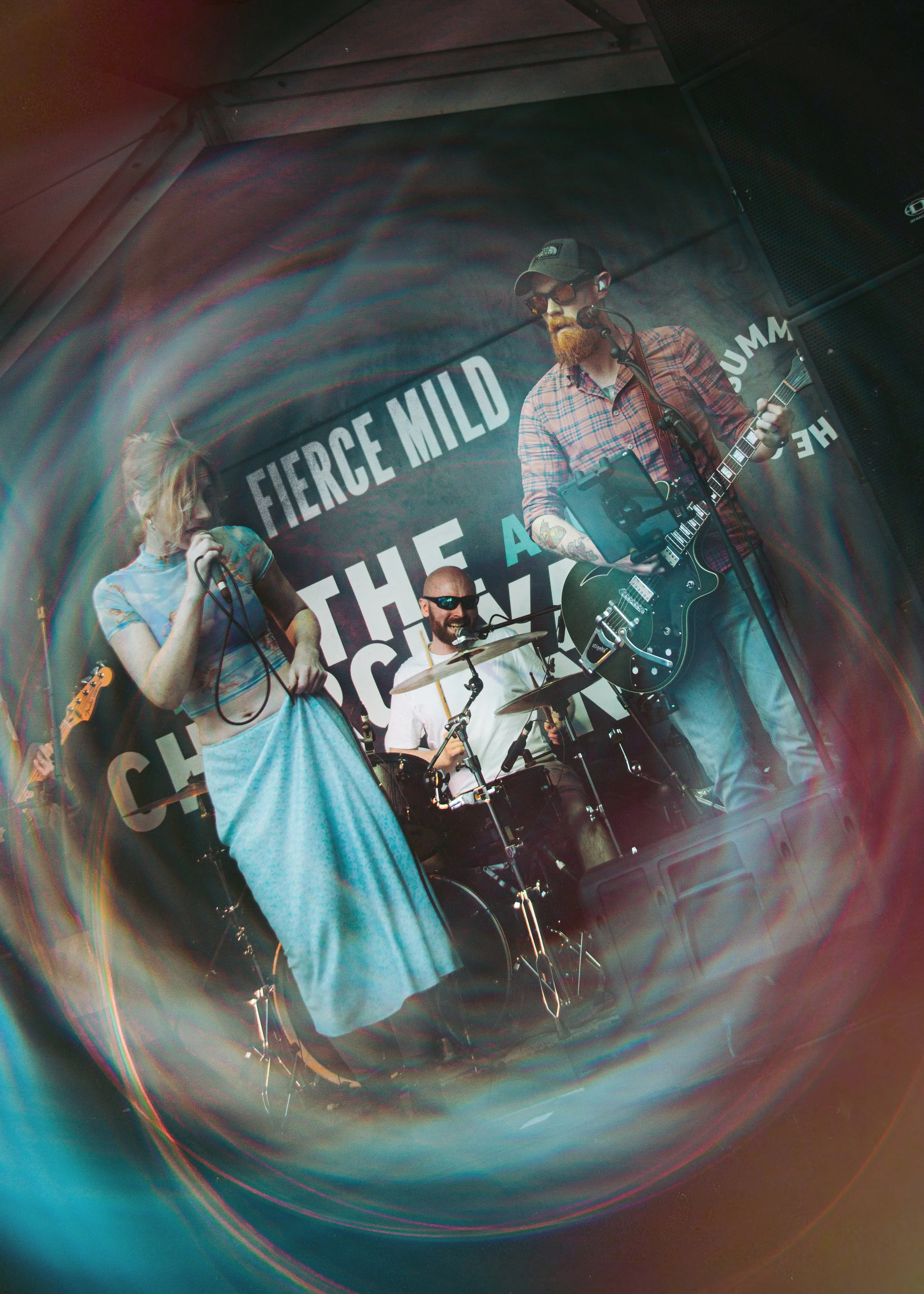 A band performing on stage with a drummer, a female singer, and a male guitarist with tattoos, sunglasses, and a cap, with a large sign in the background reading 'FIERCE MILD THE A'.