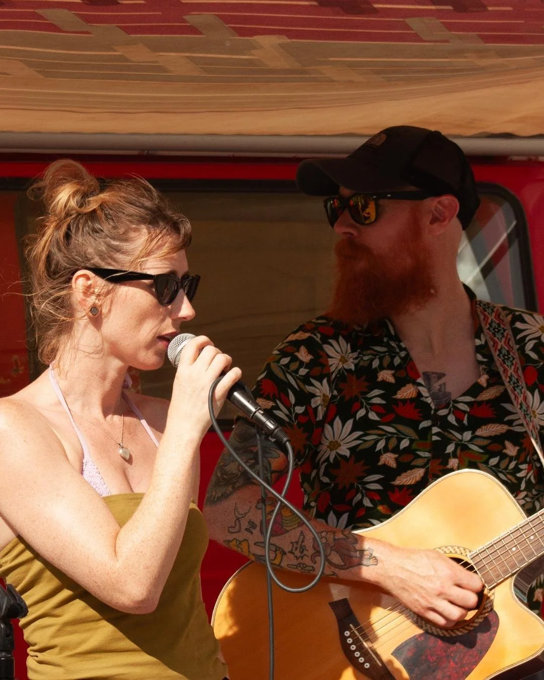 A woman in sunglasses singing into a microphone, with a man in a floral shirt, sunglasses, and cap playing guitar beside her.