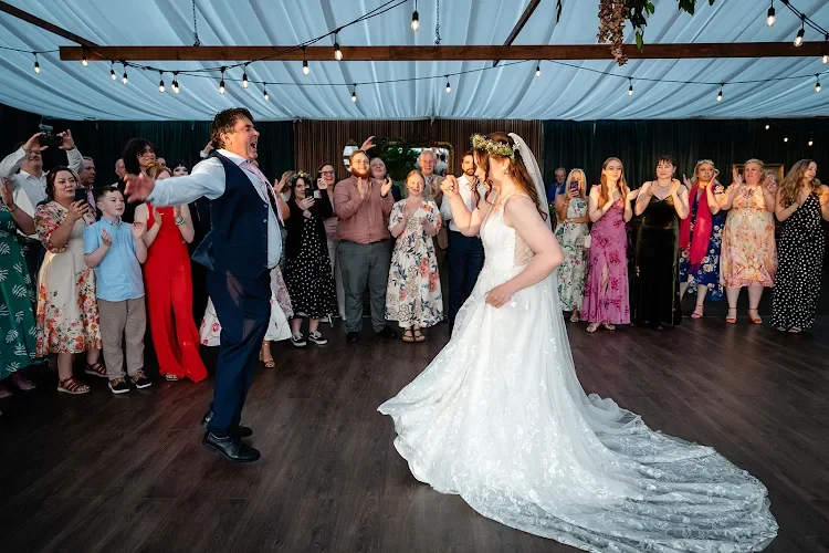 Bride and groom dancing at their wedding reception, surrounded by guests in a decorated venue with string lights.