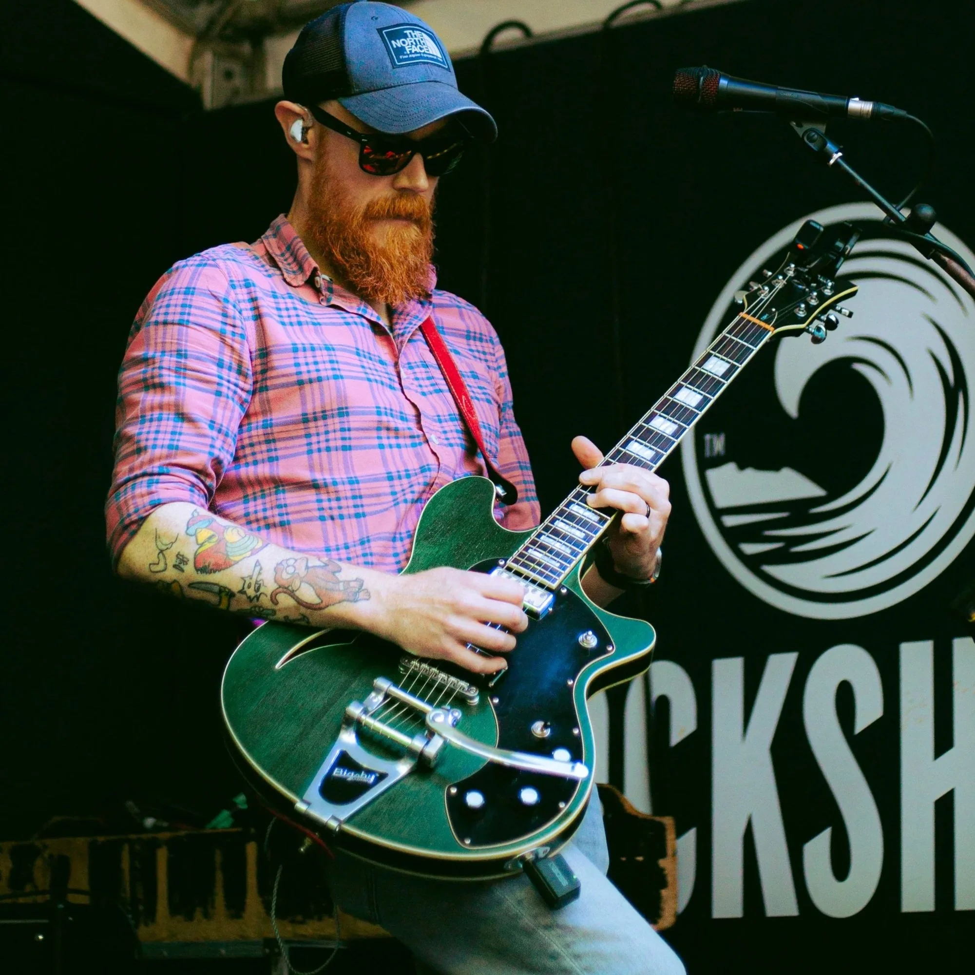 A man with a red beard wearing sunglasses, a blue baseball cap, a pink plaid shirt, and wireless earbuds, playing an electric guitar on stage with a microphone nearby.