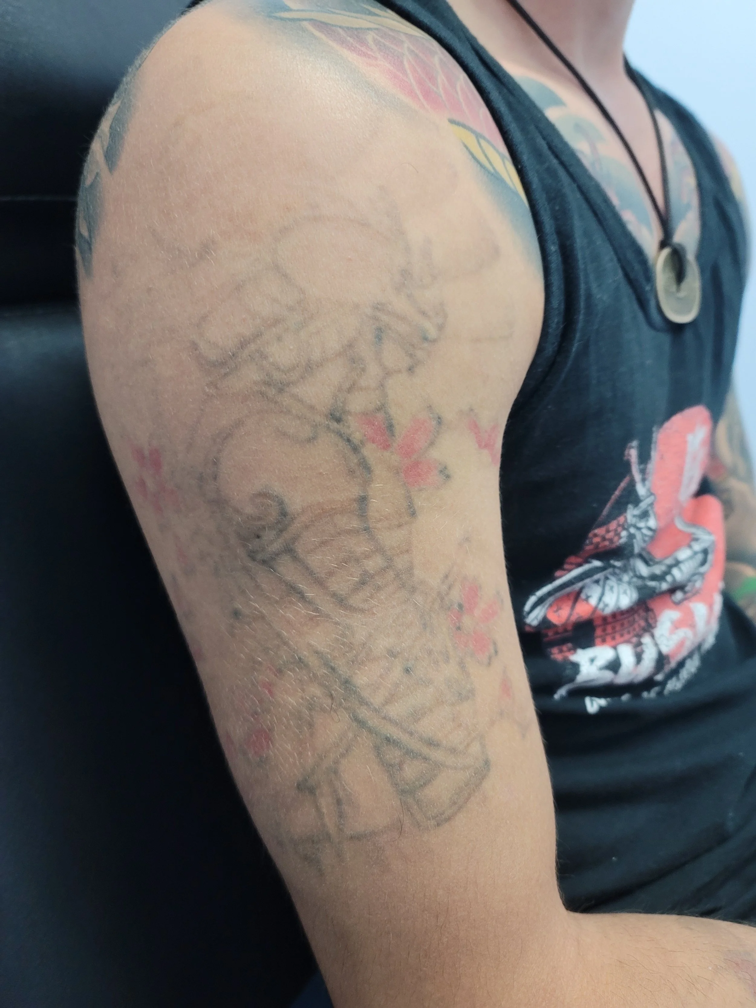 Close-up of a person's upper arm with tattoos, wearing a sleeveless black top with a graphic design.