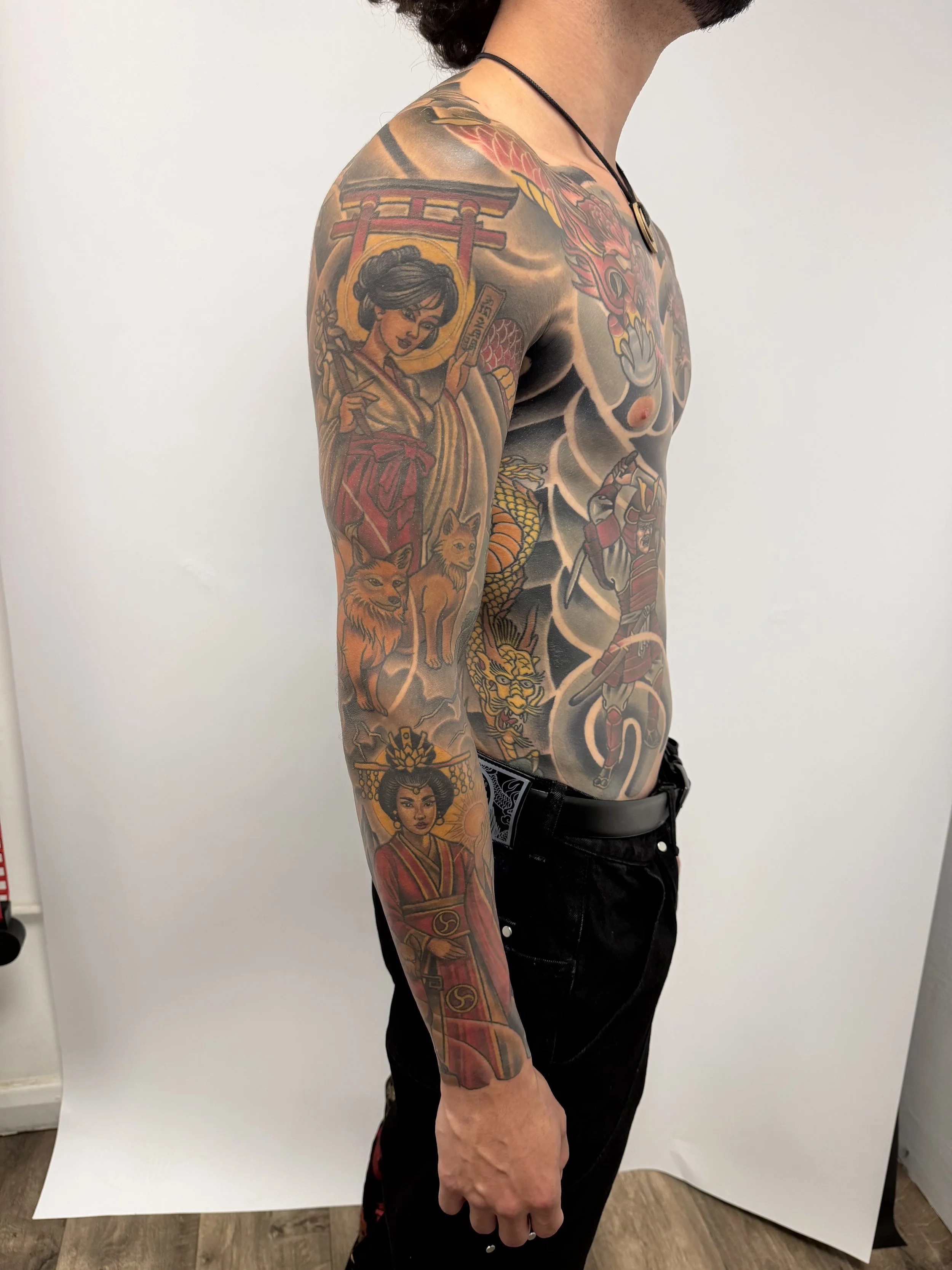 A man with tattoos covering his left arm and chest, wearing a necklace and black pants, standing against a plain white background.