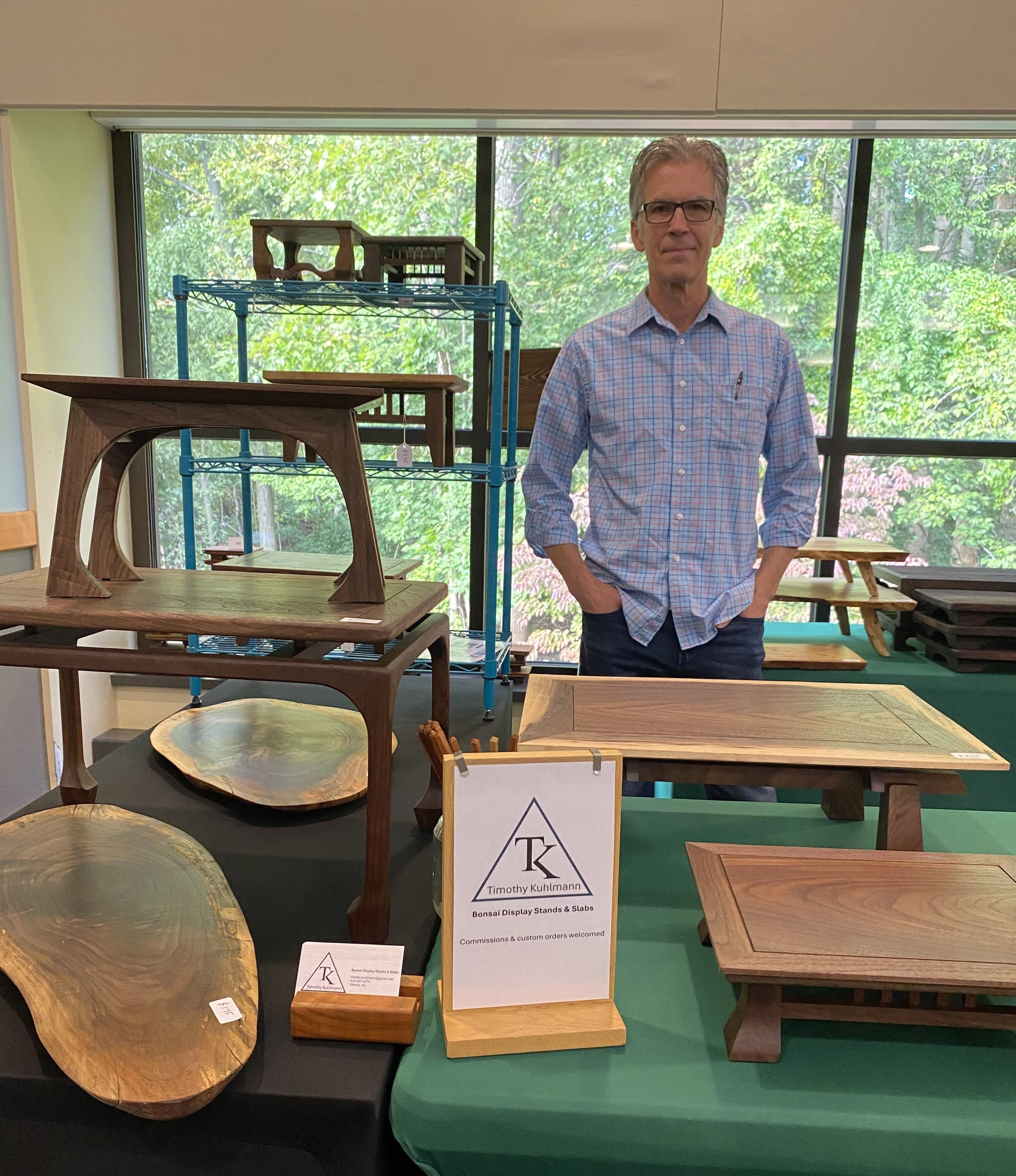 Display of various wooden bonsai display stands and slabs at a trade show, with a person standing behind the table.