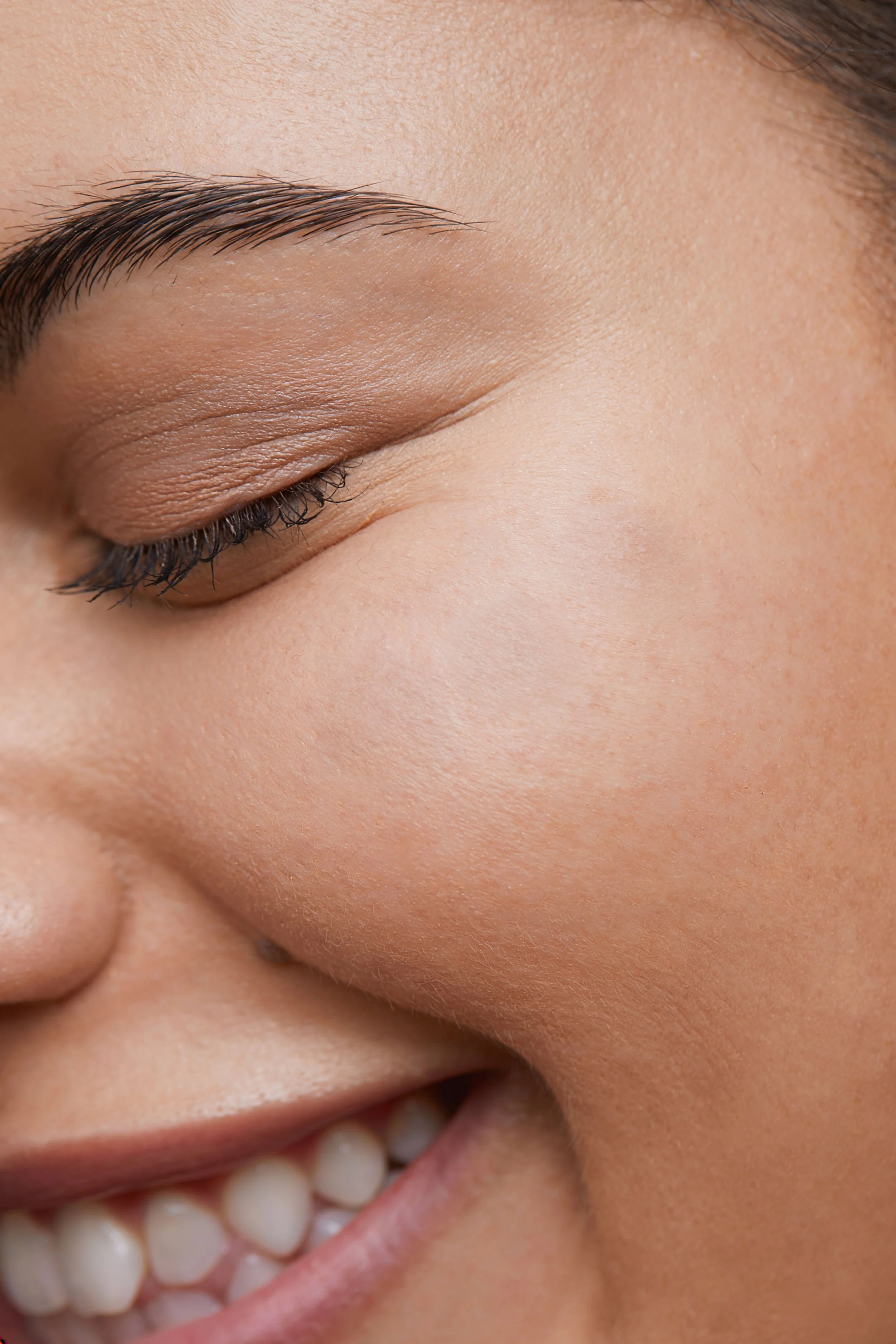 Close-up of a person's face with closed eyes, showing detailed skin texture, well-groomed eyebrows, and a partial view of a smile.