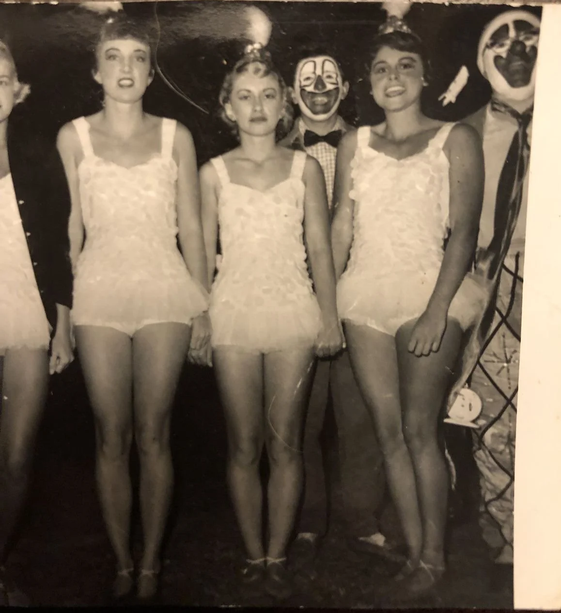 Black and white photo of four women in lace dresses standing together, with a man in a circus clown or mime costume standing behind them, wearing a painted face and a checked shirt with a bow tie.