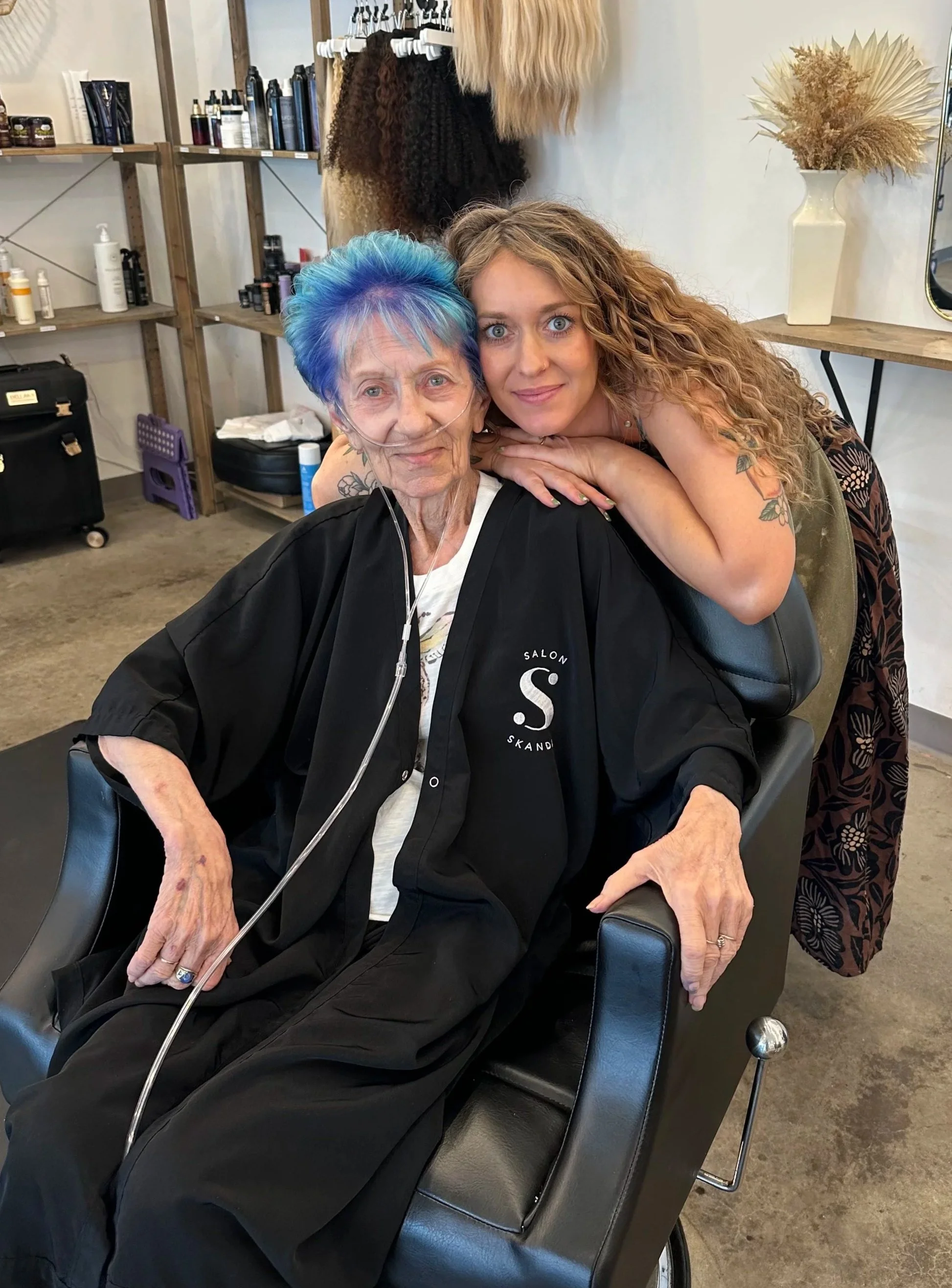 A young woman with curly blonde hair leaning over an elderly woman with short blue and purple hair seated in a salon chair. The elderly woman has a nasal oxygen tube and is wearing a black salon robe with a logo. The salon background shows shelves with hair products, a mirror, a vase with dried pampas grass, and hair extensions hanging in the background.