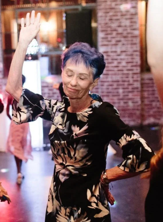 An elderly woman with short blue hair dancing with her eyes closed, raising her right hand, wearing a black and beige floral dress, in a warmly lit indoor setting with brick walls.
