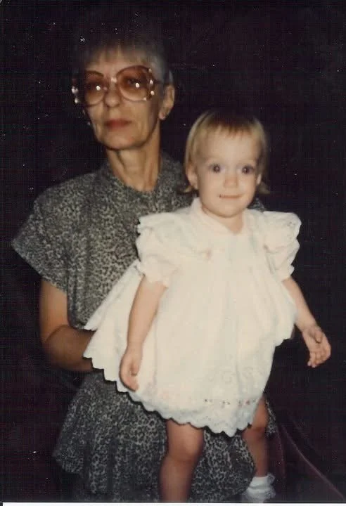 An elderly woman holding a young girl. The woman is wearing glasses and a patterned dress, while the young girl is dressed in a white dress with ruffled sleeves and a scalloped hem.