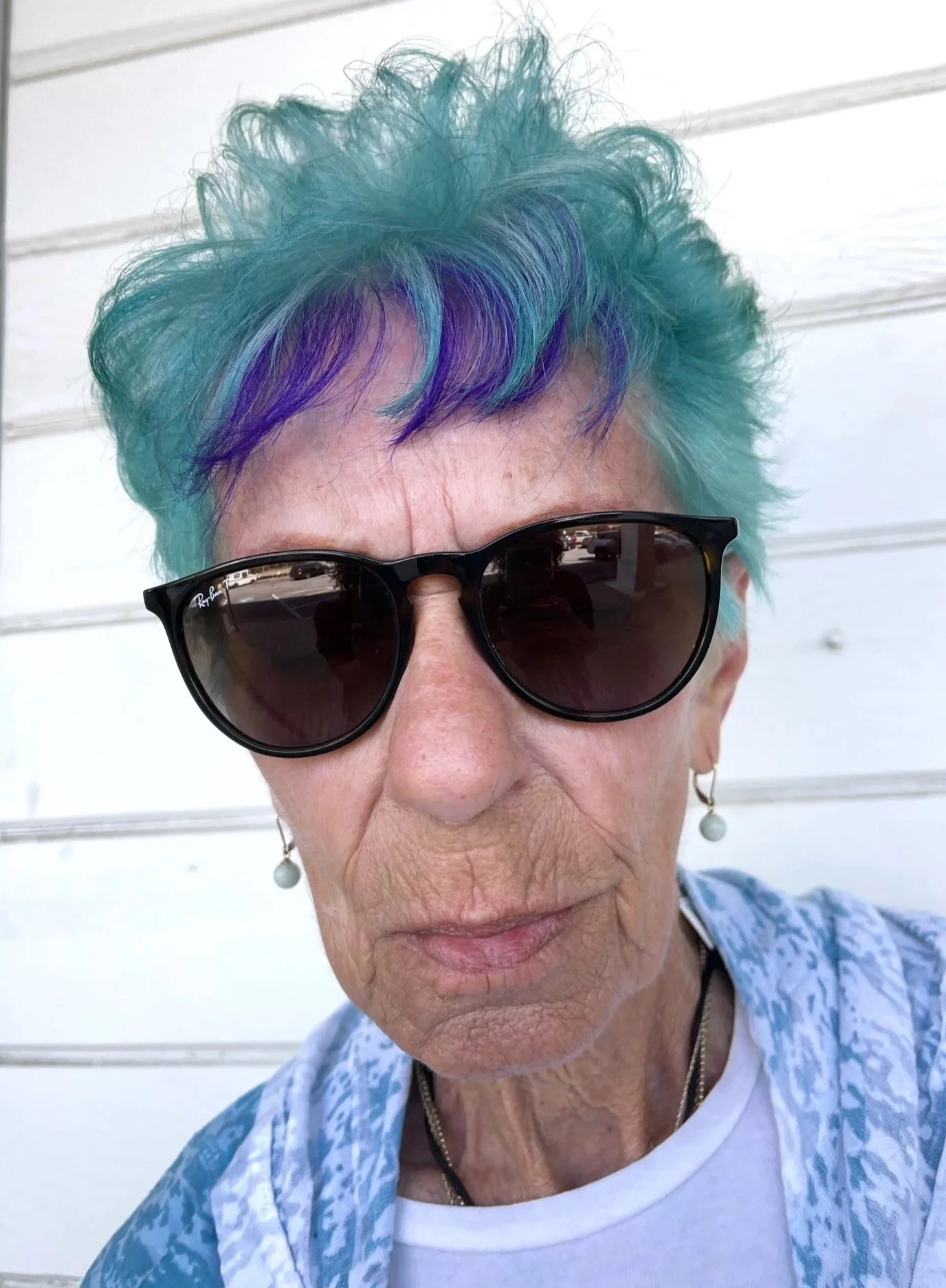 Close-up of a woman with short, curly, multi-colored hair (blue and purple), wearing black sunglasses, pearl earrings, and a blue patterned shirt. She is outside in front of a white wall.
