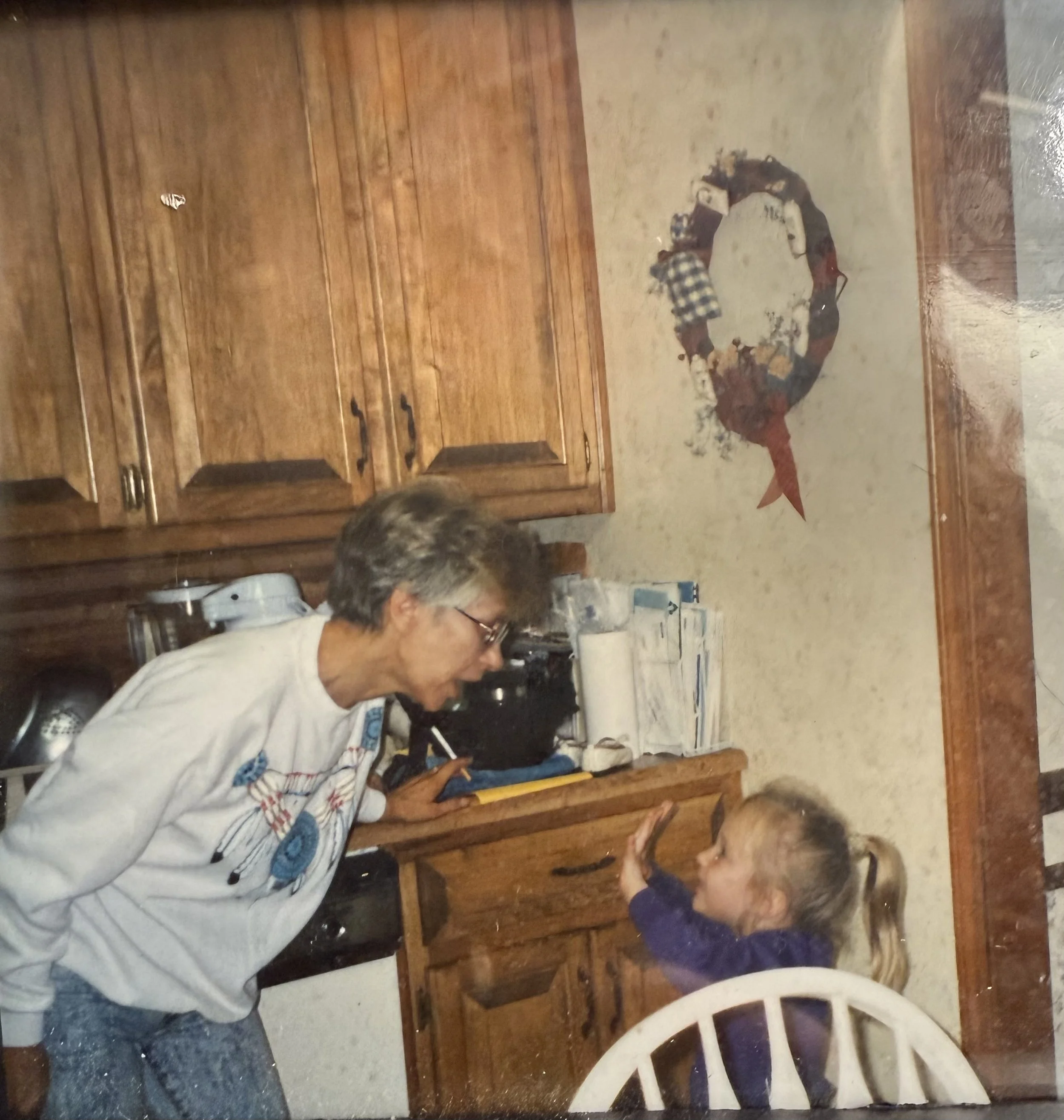 A woman with glasses and short hair, wearing a gray shirt, is sitting at a dining table with a child with long hair sitting across from her. They are playing a game involving a circular board with pieces and tokens on it. The room has wooden cabinets and a tiled floor.