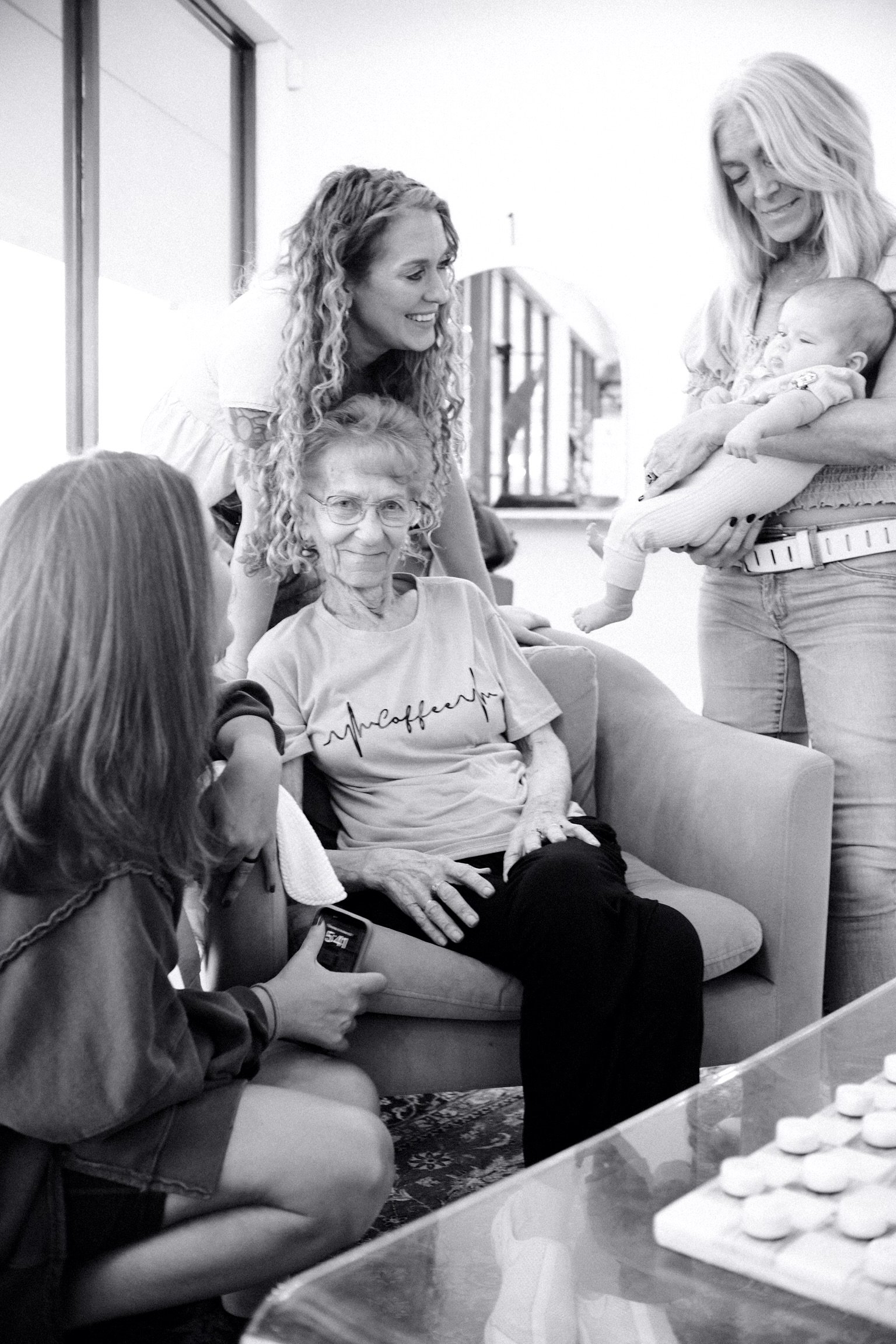 A group of women and a baby sitting inside a living room, engaging in a conversation and smiling. The woman holding the baby is smiling and looking at the baby, while the woman standing is leaning over and smiling at them. A woman in the foreground is using her phone.