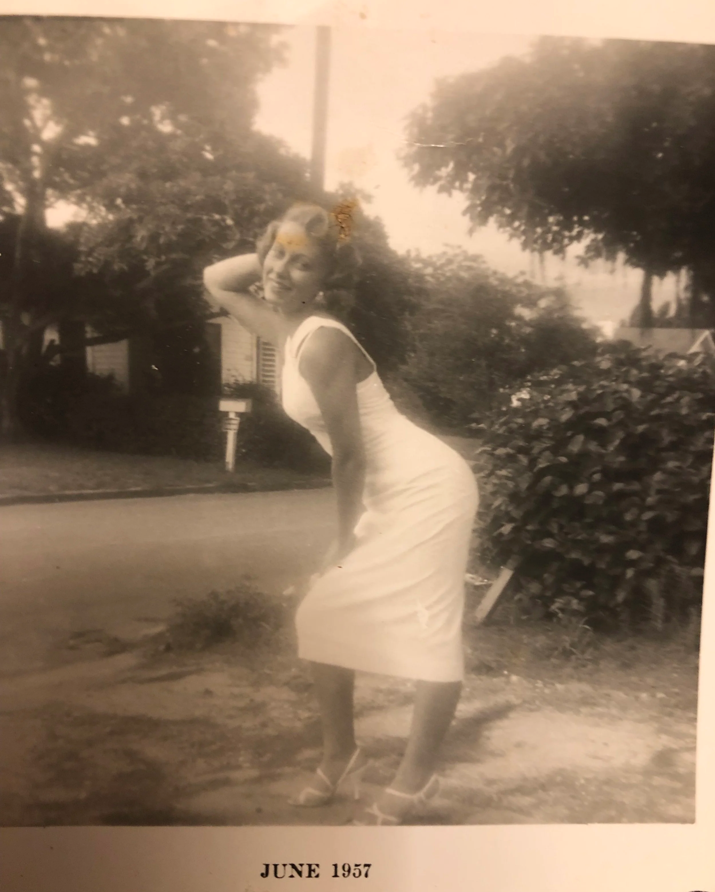Black and white photograph of a woman smiling outdoors, wearing a strapless dress with a ribbon, in June 1957, surrounded by trees and a house.