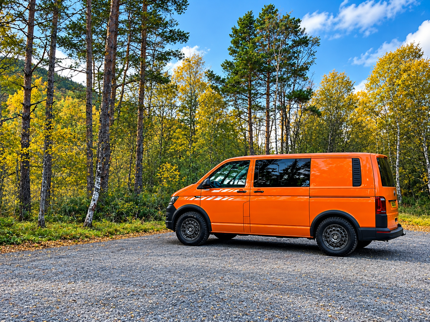 Orange camper van parked on a gravel road in a forest with green and yellow trees under a blue sky.