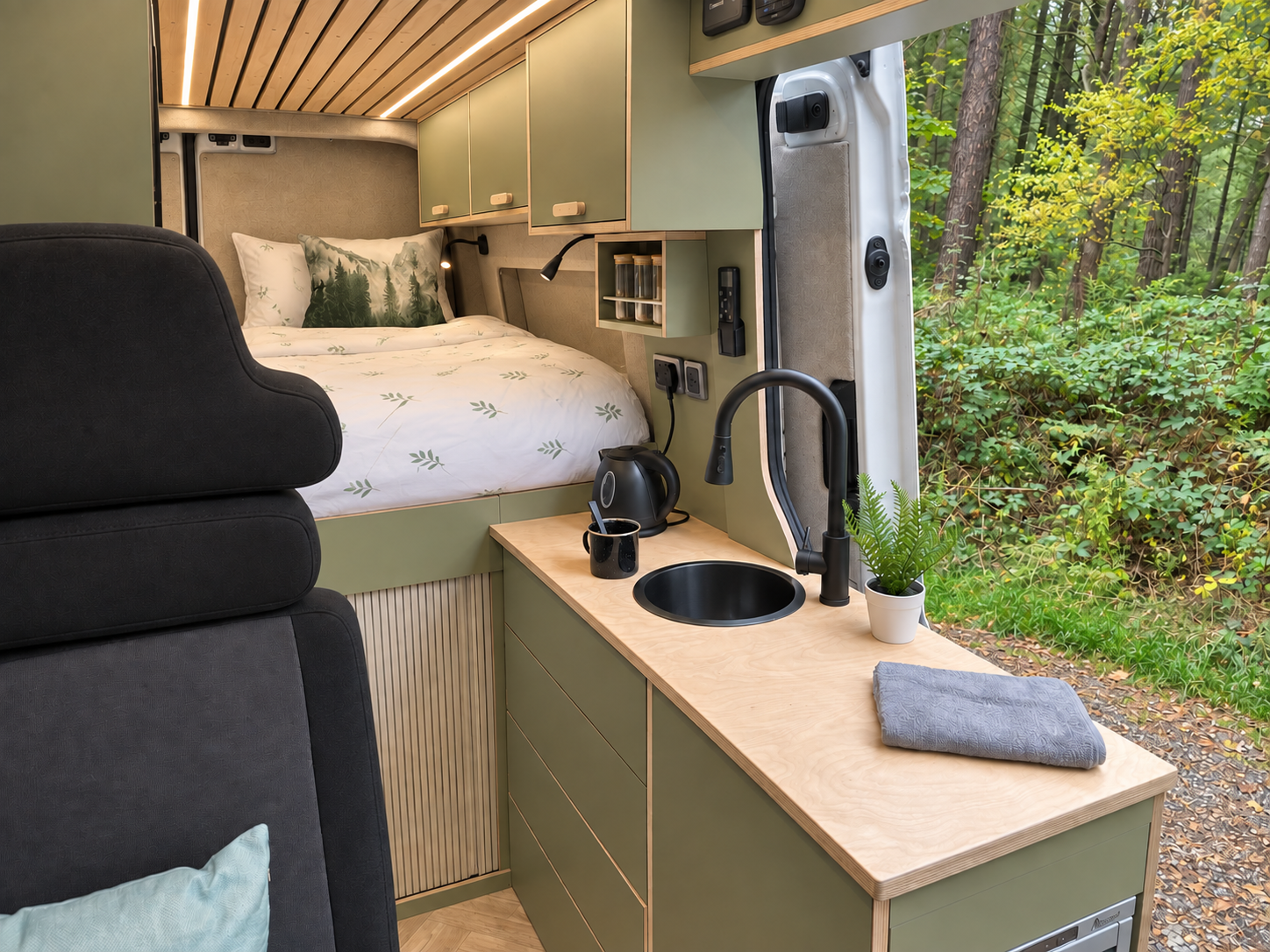Interior of a small camper van with a bed, black armchair, small kitchen counter with sink, electric kettle, mug, and a potted fern, with a window showing green trees outside.