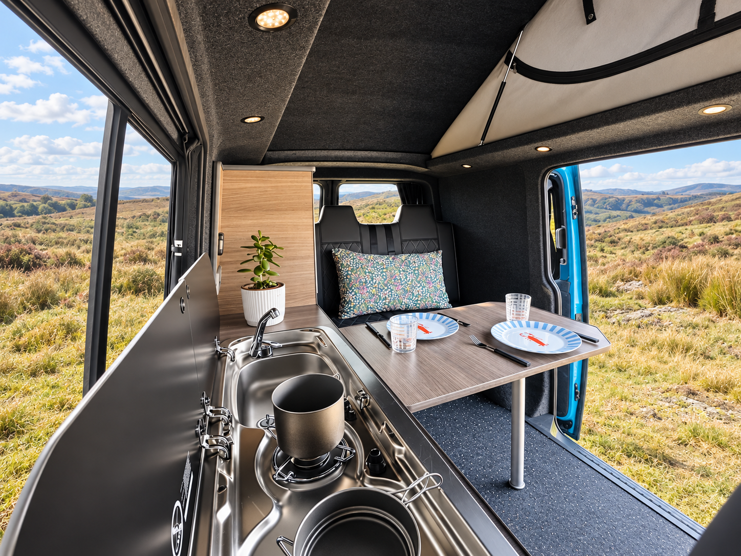 Interior of a camper van with a small kitchen area, a table set with plates, glasses, and utensils, and a window showcasing a scenic outdoor landscape of grassy fields and rolling hills.