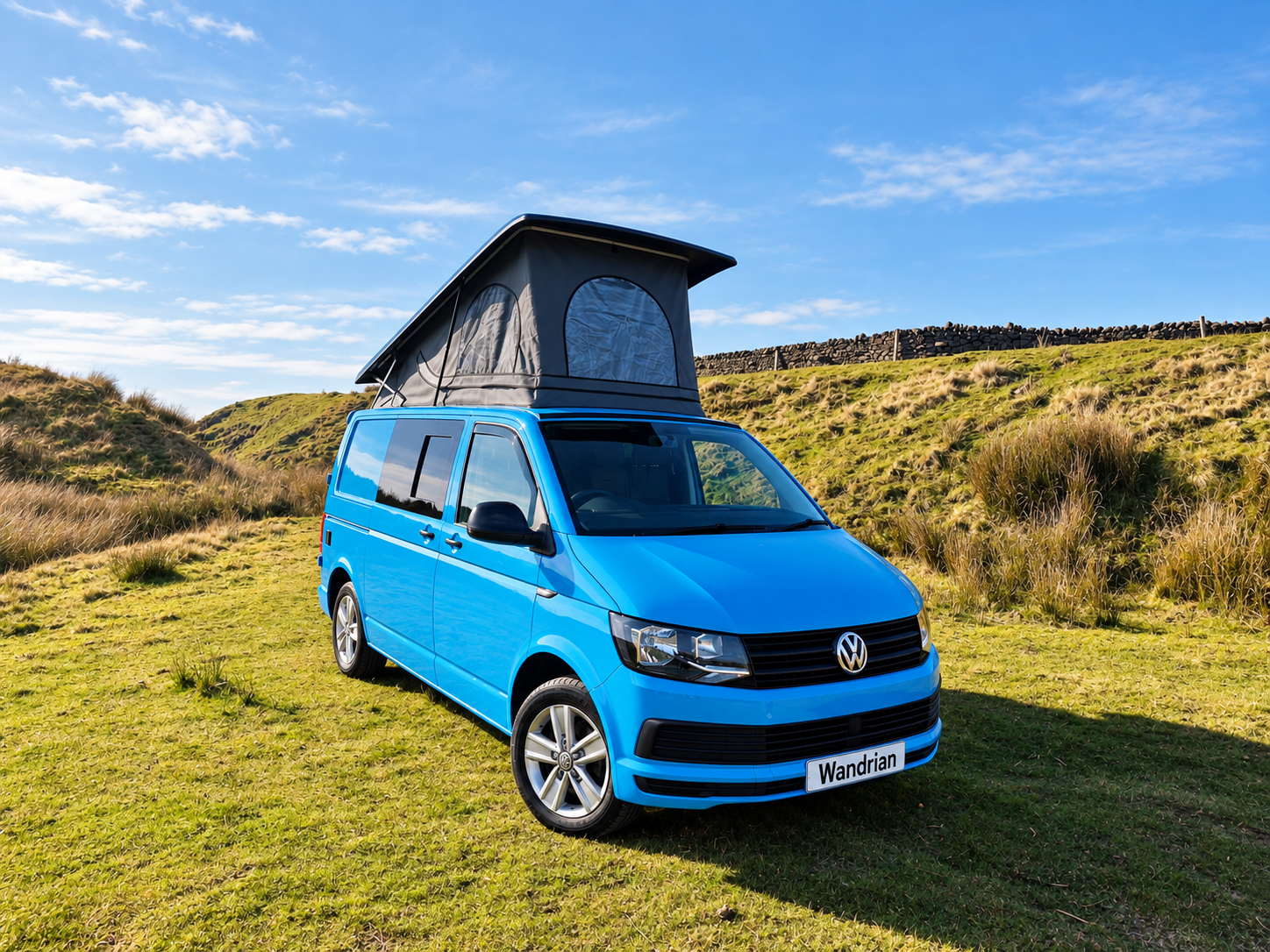 A blue Volkswagen camper van with a pop-top roof parked on grassy terrain surrounded by hills under a partly cloudy sky.