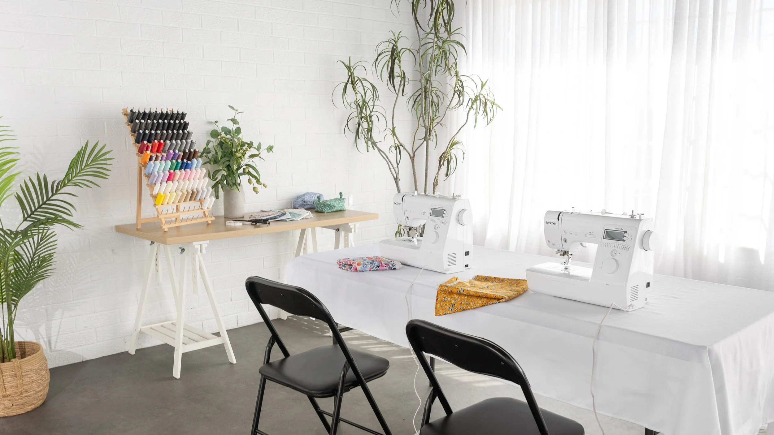 A craft room with two sewing machines on a white table, a basket with plants nearby, a table with thread spools and sewing supplies, and a white brick wall with a large potted plant.