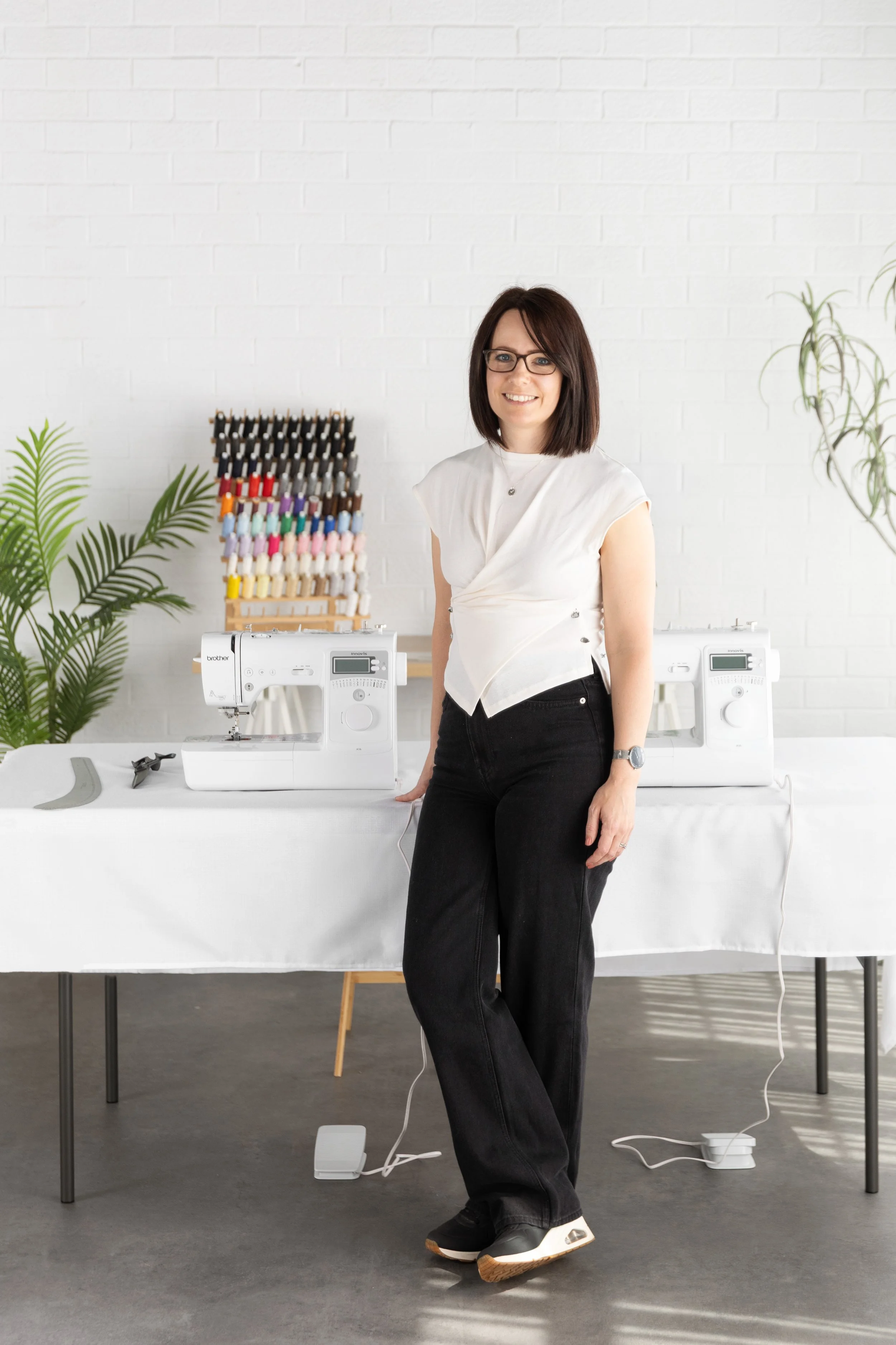 A woman with dark hair, glasses, and a white sleeveless top standing next to two sewing machines, with spools of thread and green plants in a brightly lit, modern room with a white brick wall.