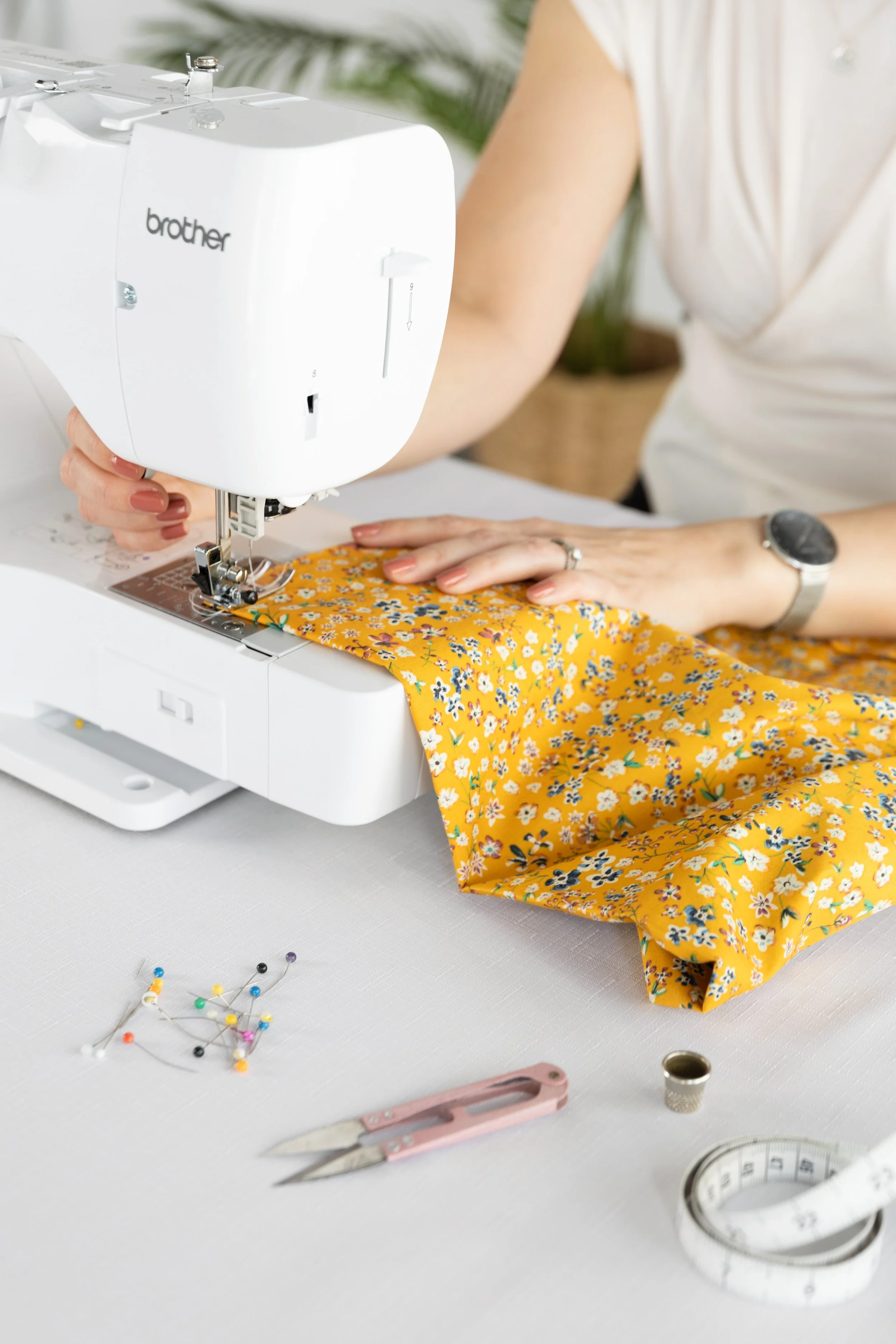 A person sewing with a Brother sewing machine on a white table. They are wearing a white top and a watch on their wrist. Tools and sewing pins are on the table, and a yellow fabric with a floral pattern is under the sewing machine.