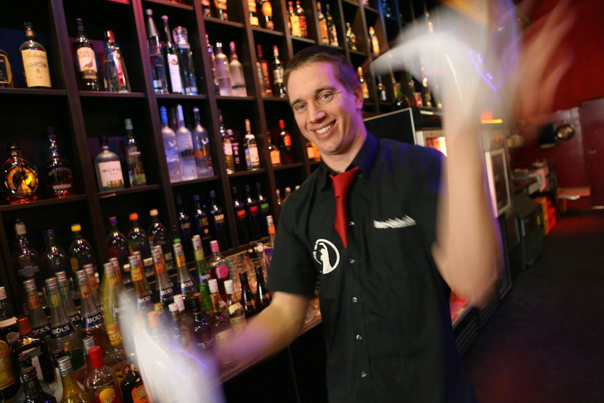 A smiling bartender, dressed in a black shirt with a red tie, stands behind a bar stocked with various bottles of alcohol, with a rack of spirits in the background.