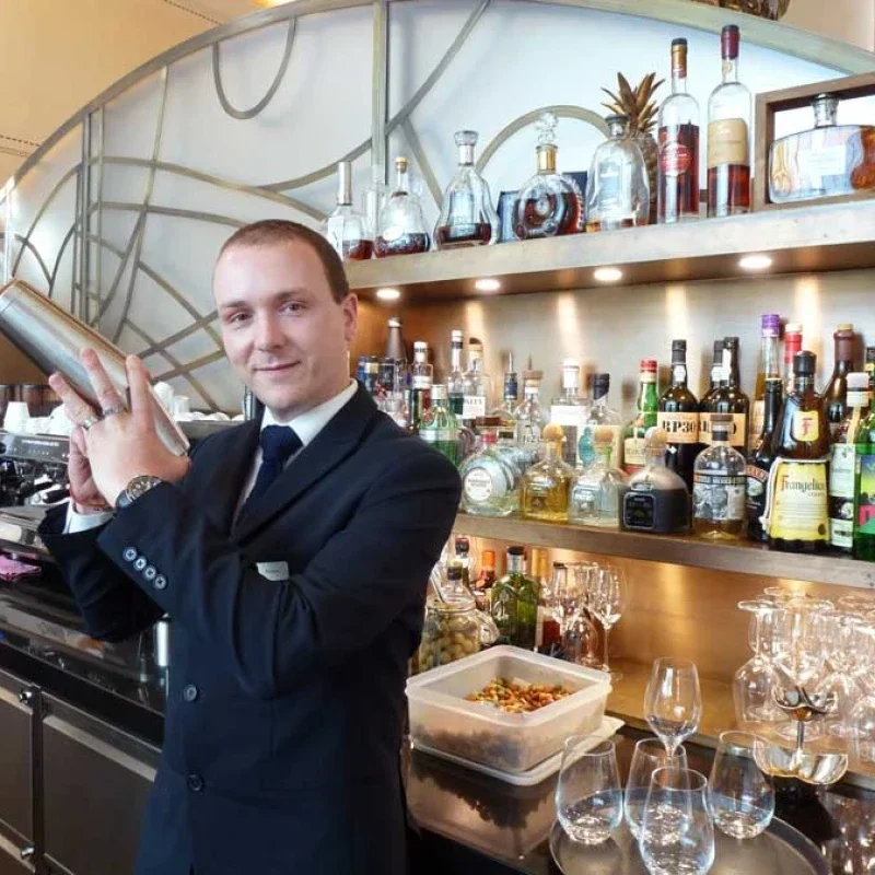 A bartender in a dark suit holding a cocktail shaker in a bar with alcohol bottles on shelves behind him