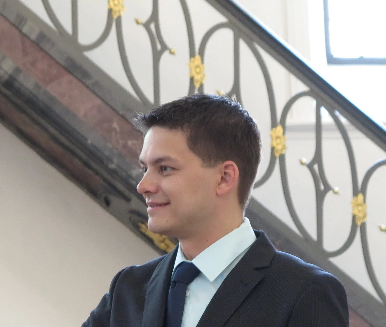 A young man with short dark hair in a suit and tie standing in front of a decorative staircase railing with yellow floral designs.