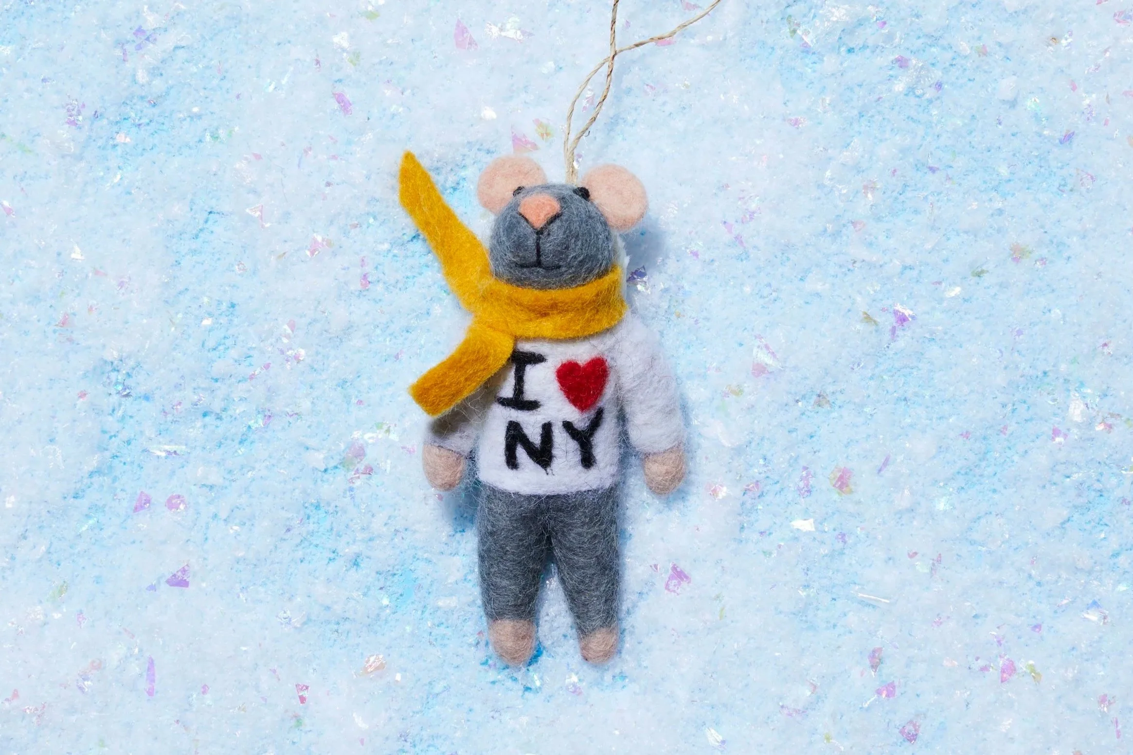 Handmade felt mouse ornament wearing a yellow scarf and white “I ♥ NY” shirt on a blue glitter background