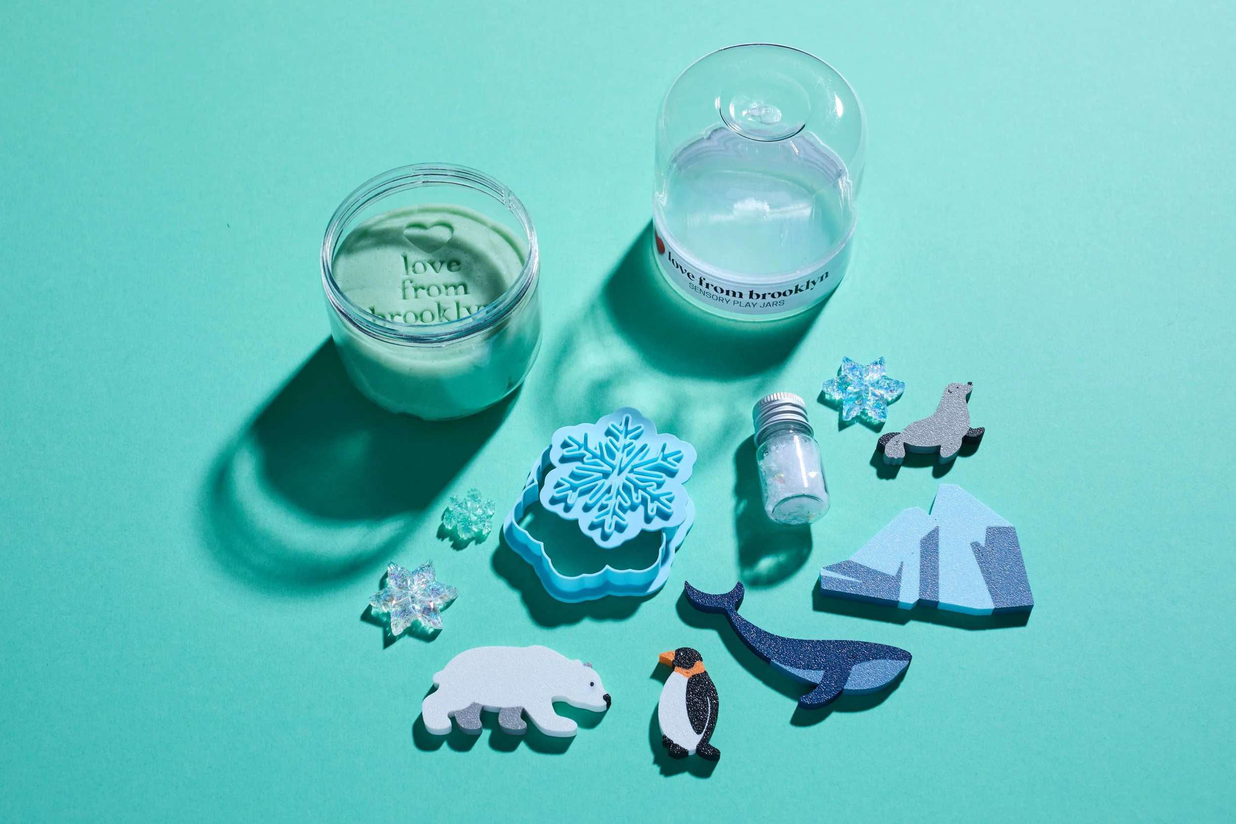 Sensory play jars with green modeling dough, snowflake molds, glitter, and polar-themed wooden figures on a turquoise background