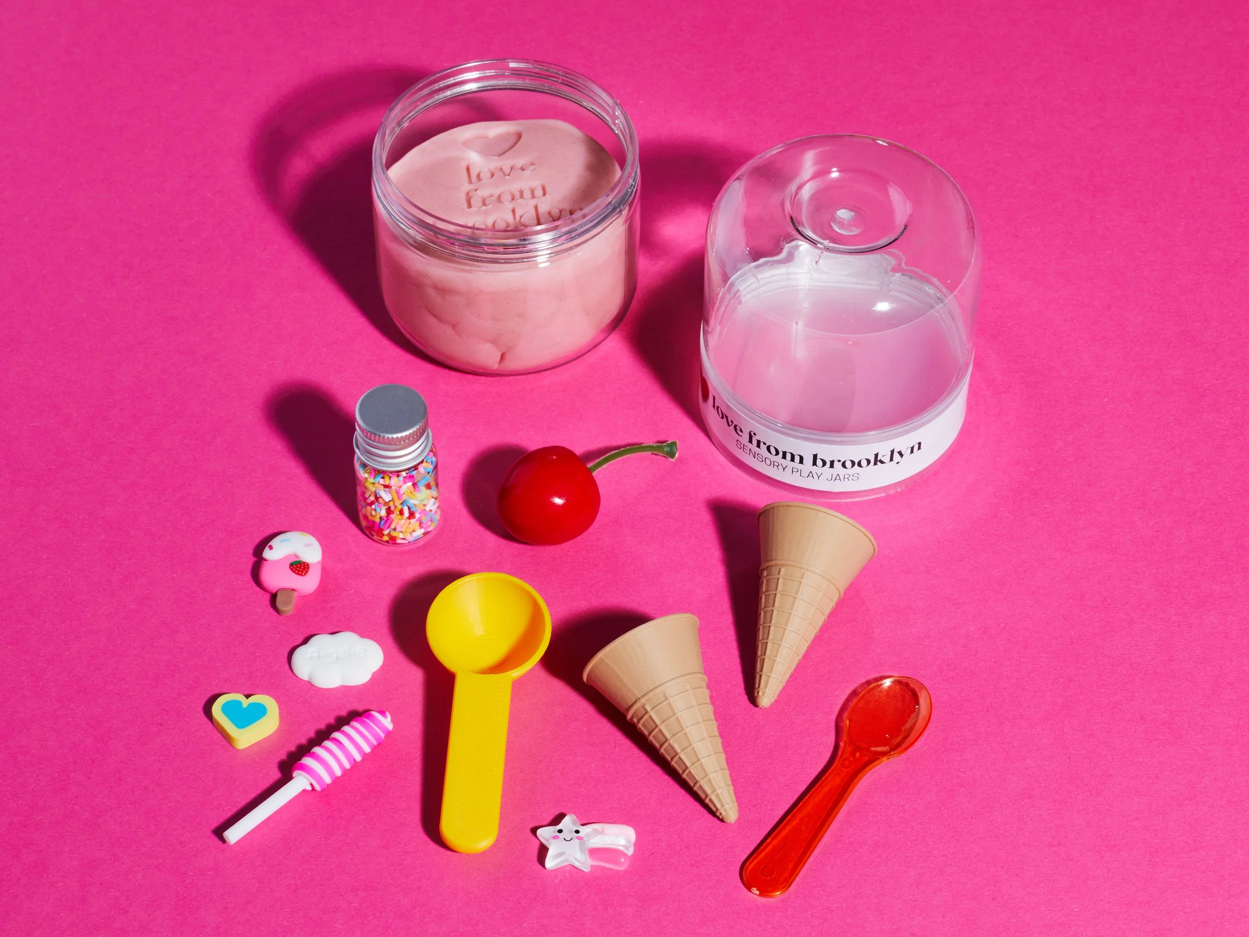 Pink sensory play setup with homemade play dough, ice cream cones, colorful sprinkles, toy fruit, scoops, spoons, and a candle jar on a bright pink background