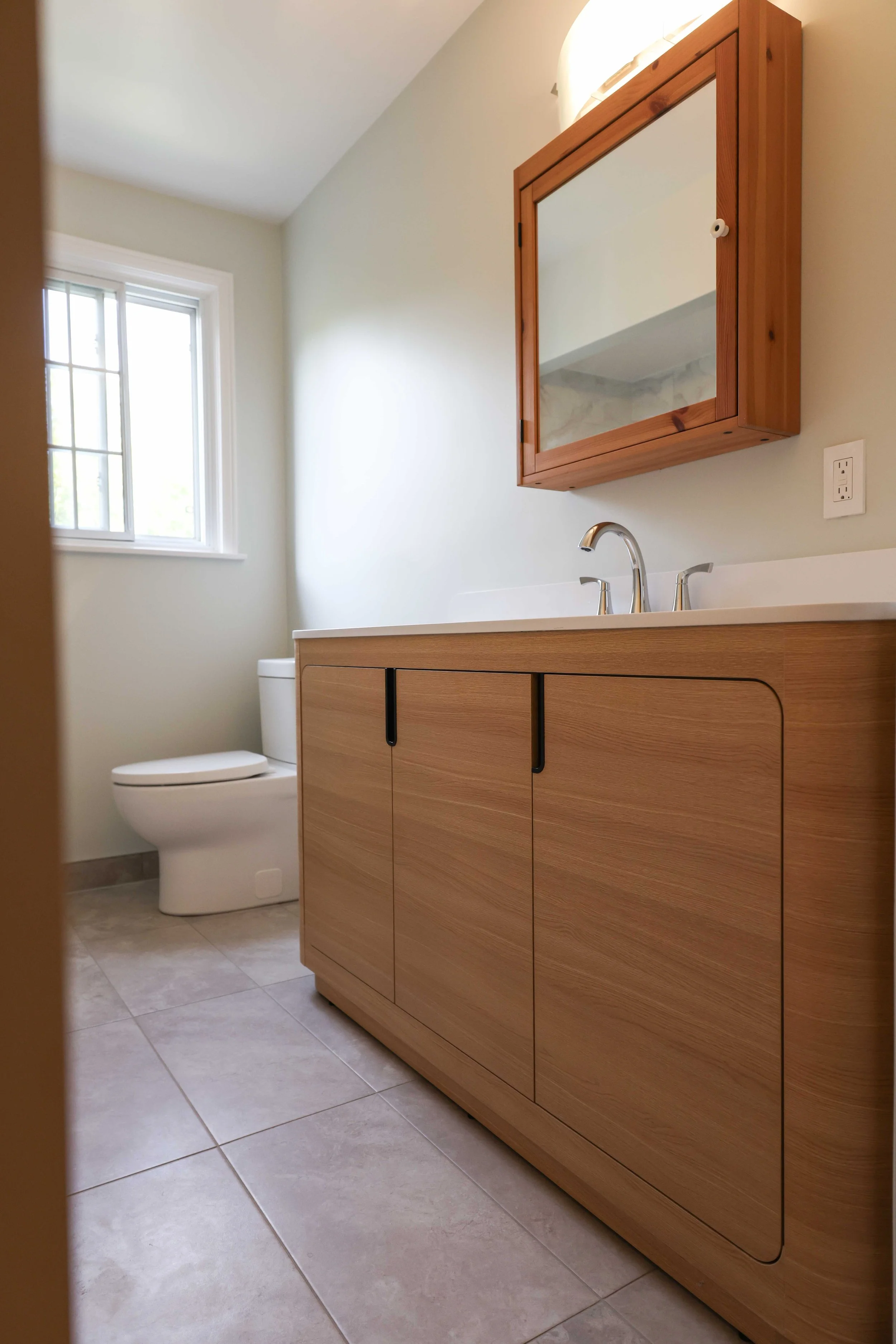 A bathroom corner with a toilet, a wooden vanity with a white countertop and a built-in sink, a mirror cabinet above the sink, a small window with white trim, and tiled flooring.