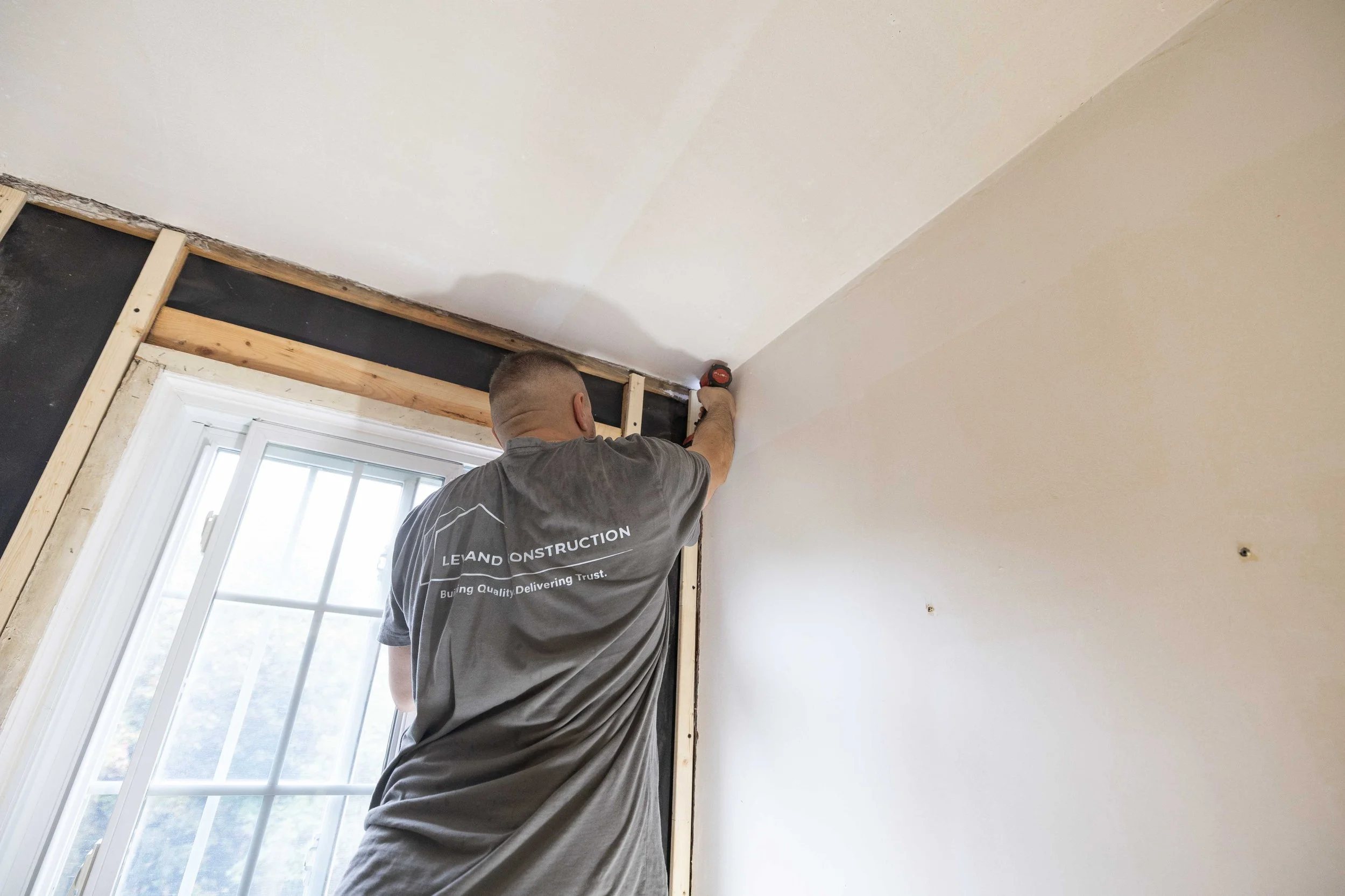 Man installing drywall near a window with power tools, in a room under renovation.