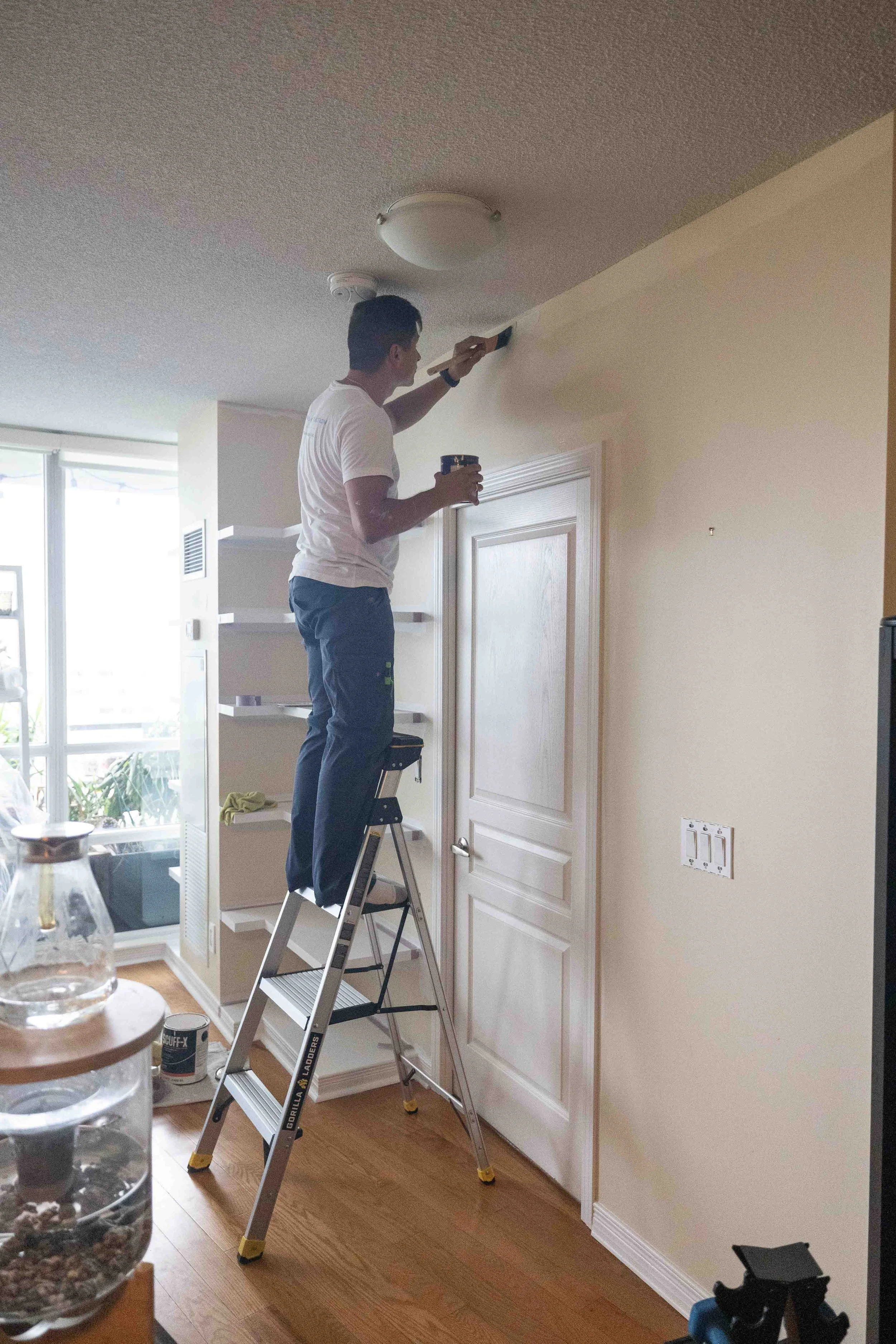 A man on a step ladder using a putty knife to repair or paint the corner of a ceiling in a living room. The room features a door, an electrical outlet, and a window with sunlight coming through.