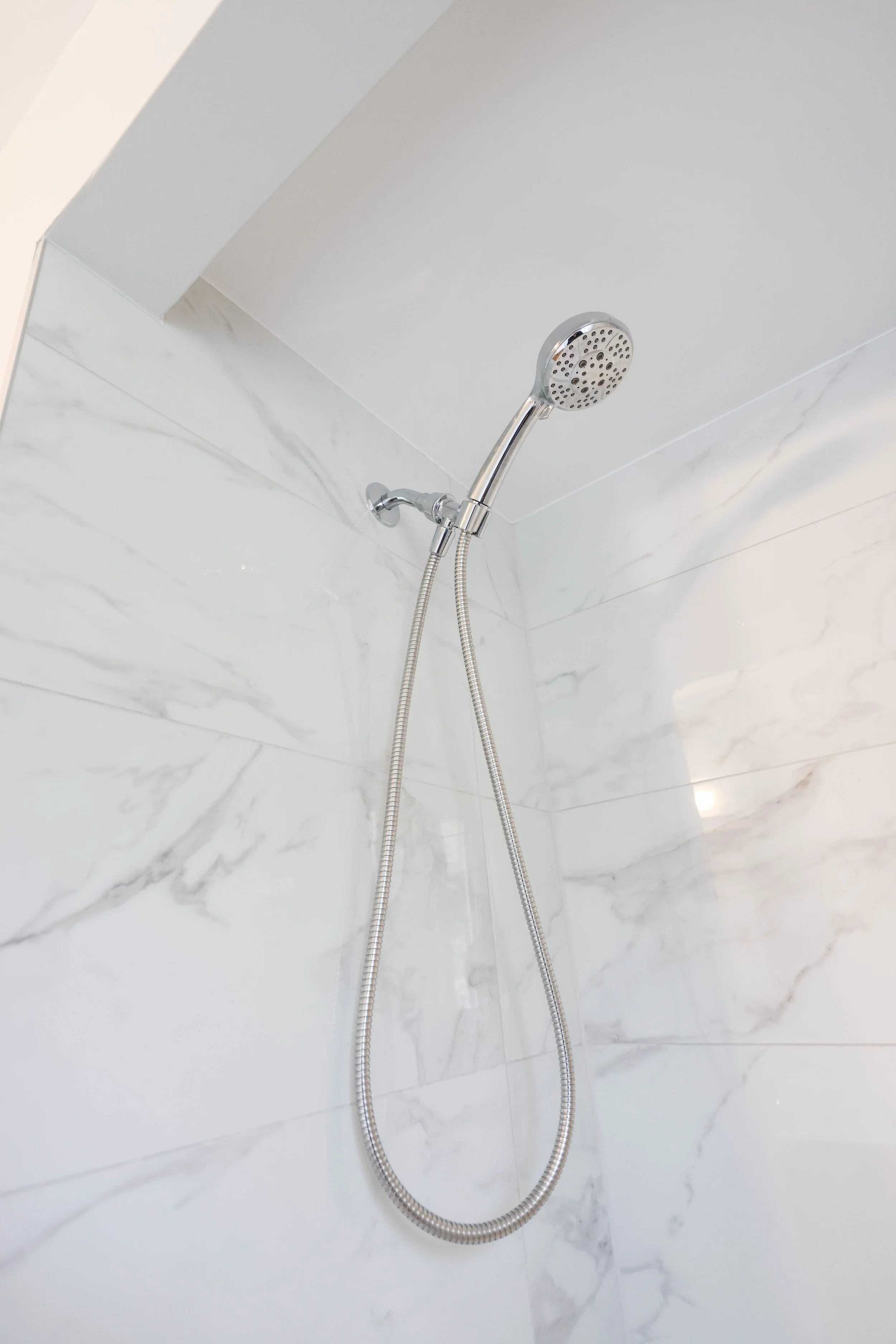 A chrome handheld showerhead mounted on a white wall with marble pattern, connected to a flexible hose.
