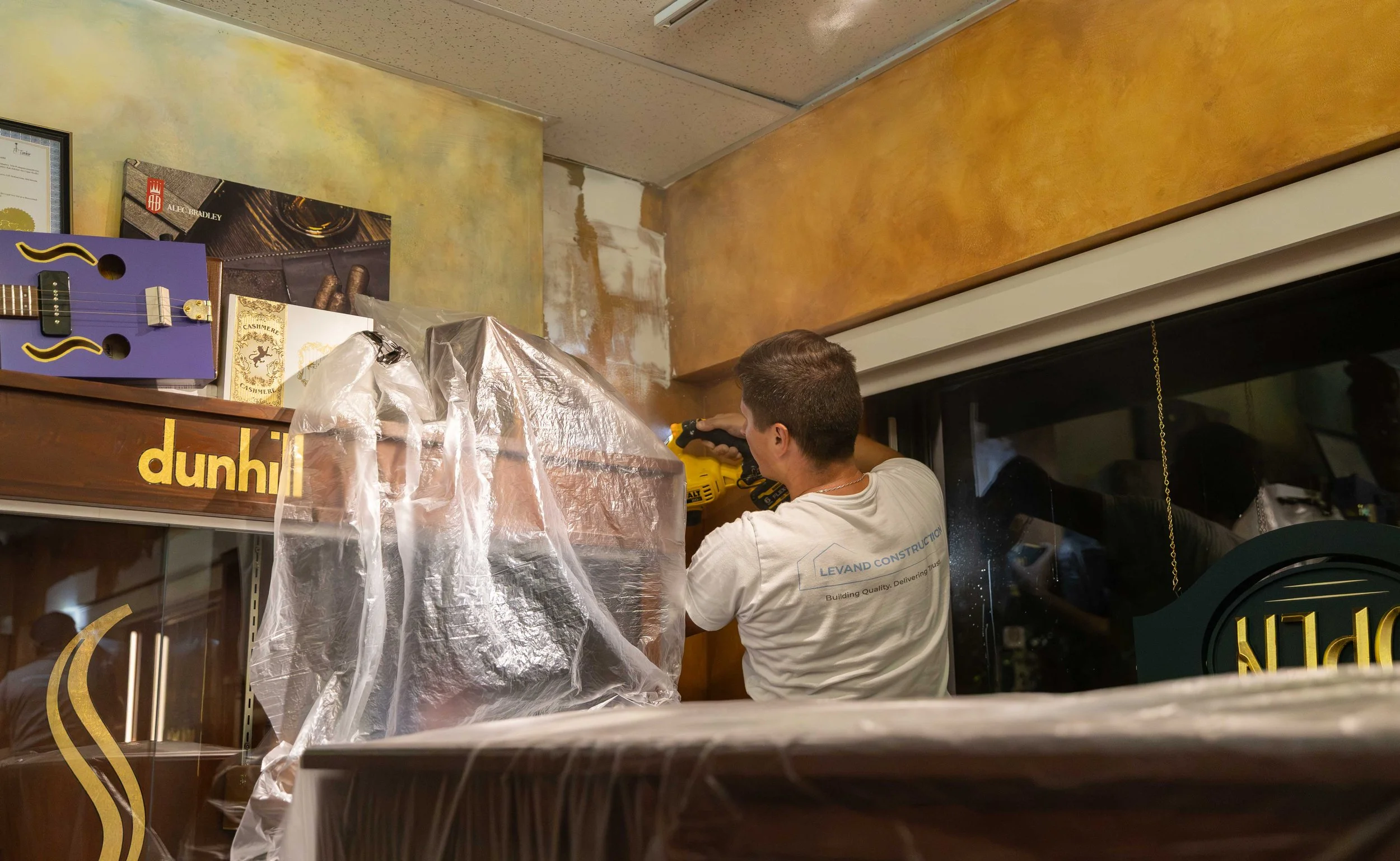 A man working on a ceiling with tools, standing next to a window and a wall with various posters and signs.