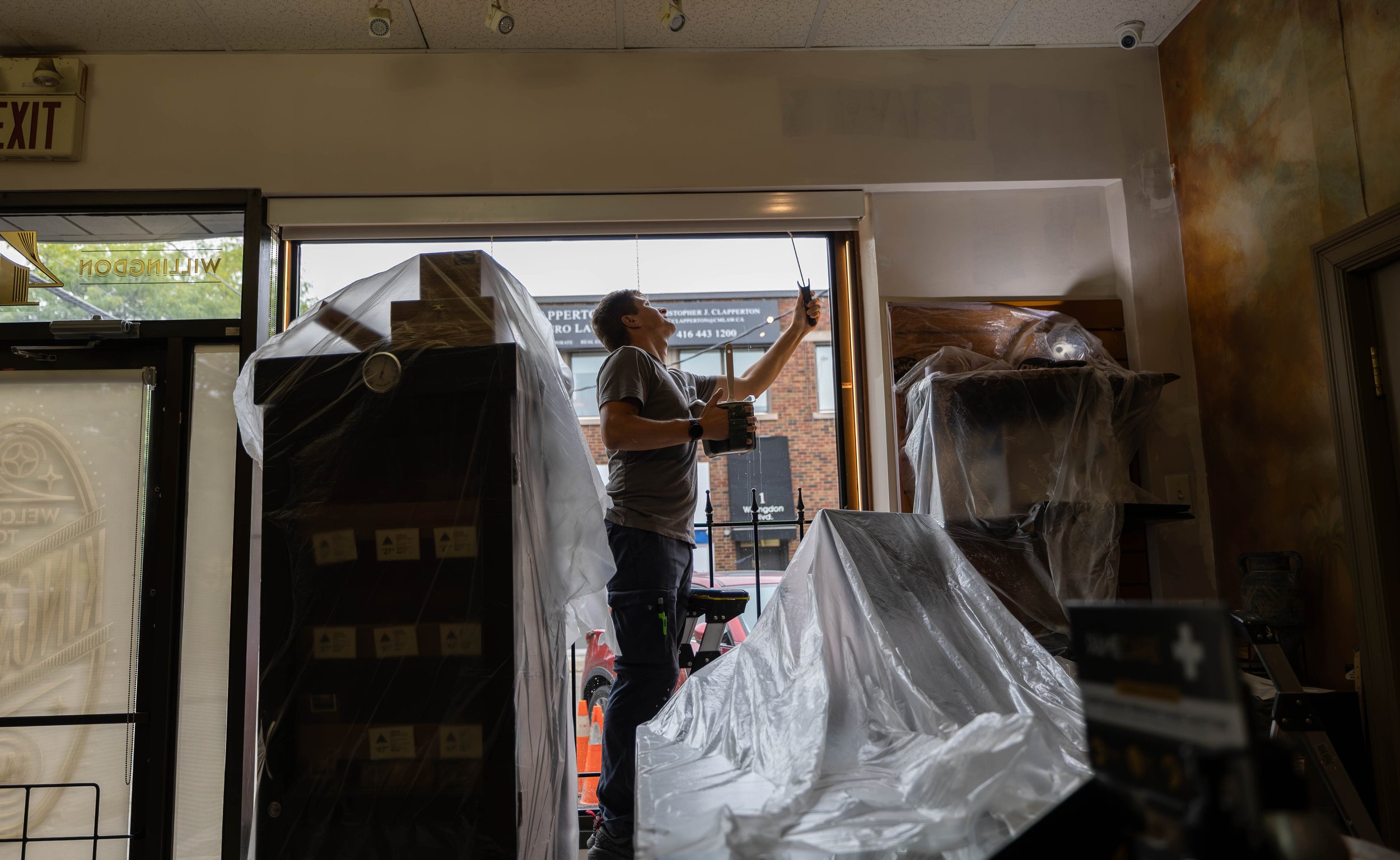 A man standing inside a store near the entrance, holding a remote-controlled device in one hand and a cup in the other, with shelves and large furniture covered in plastic around him, and a window showing the street outside.