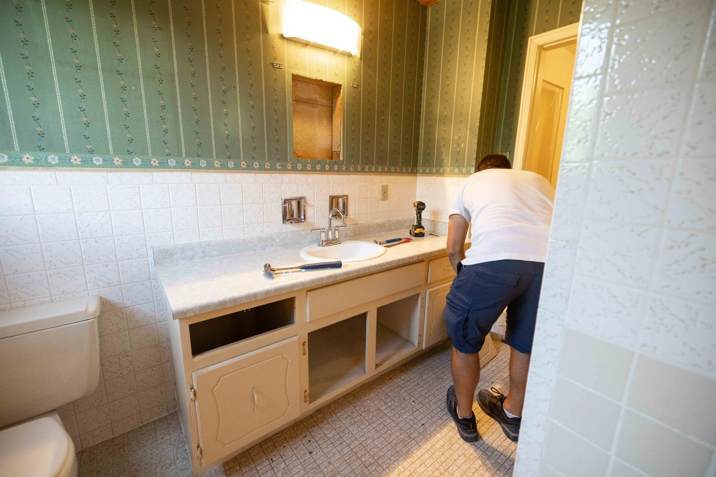 A person working on a bathroom vanity with tools, removing the cabinet doors, in a bathroom with green patterned wallpaper and white tiled walls.