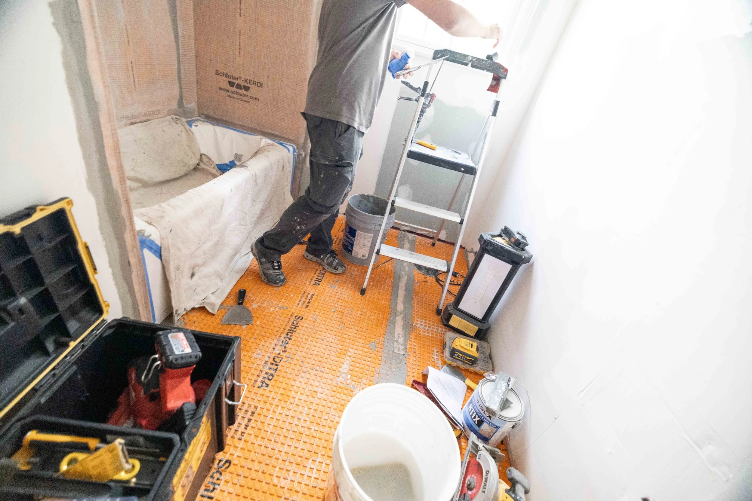 A construction worker stands inside a room with unfinished walls. He is holding a small tool in his right hand and facing a window. There are various tools and containers around, including a black toolbox, a yellow drill, a ladder, and paint buckets.