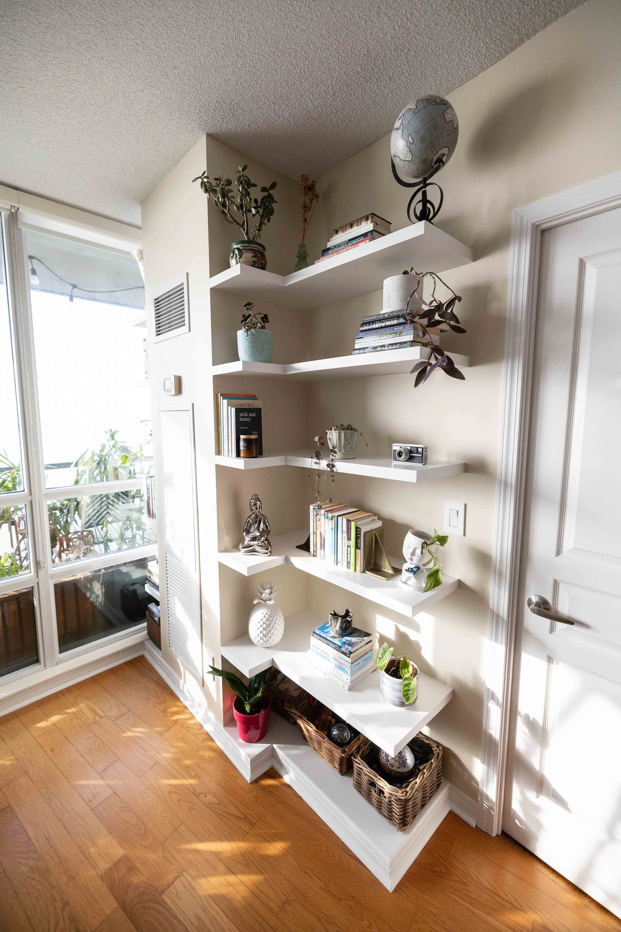 A white wall with five floating shelves holding books, plants, and decorative items. To the left, a glass sliding door with a balcony and outdoor furniture. The floor is wooden.