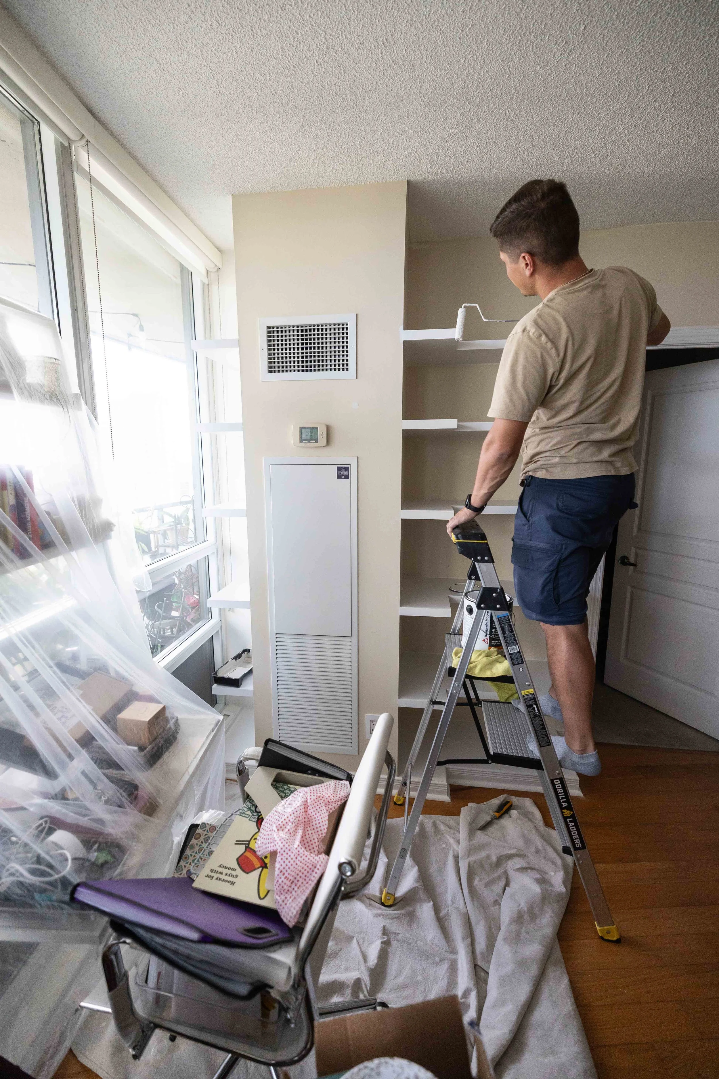 A man standing on a step ladder painting the top shelf of a white bookshelf in a bright living room with large windows.